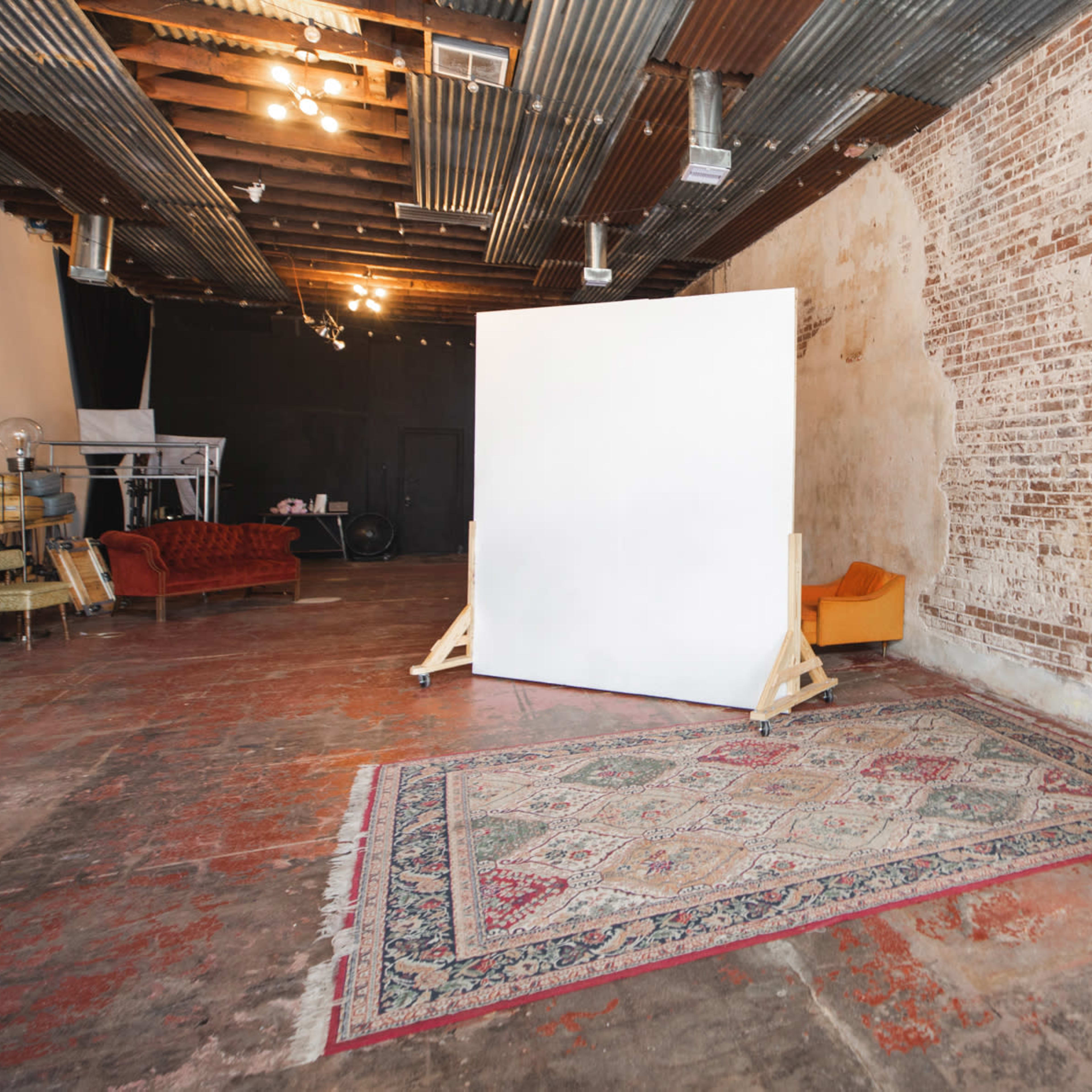 An industrial-style studio with exposed brick walls, a large white backdrop, and a colorful area rug on a concrete floor.