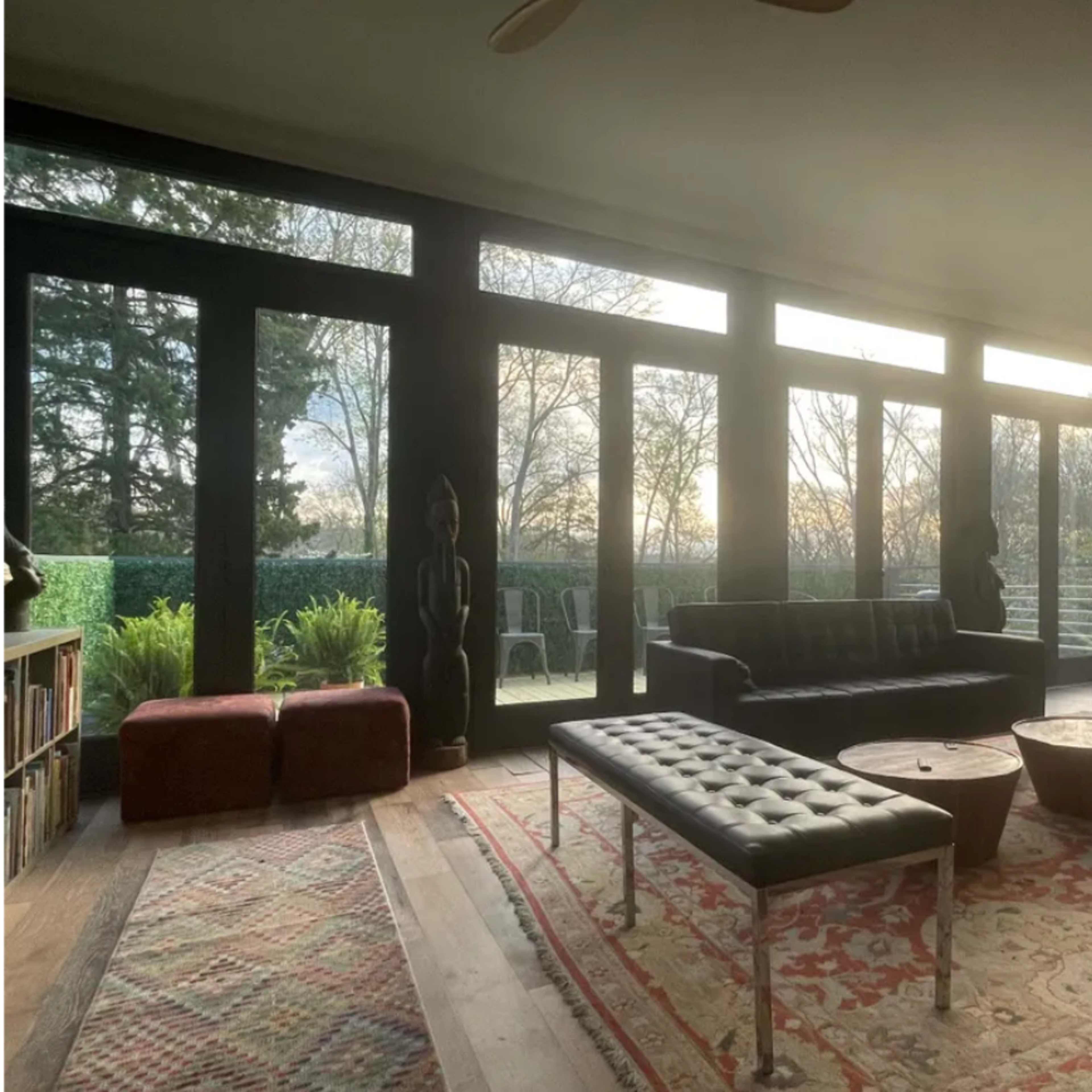 A sunlit living space with large windows, featuring a black sofa, a wooden coffee table, and a vintage motorcycle visible outside.