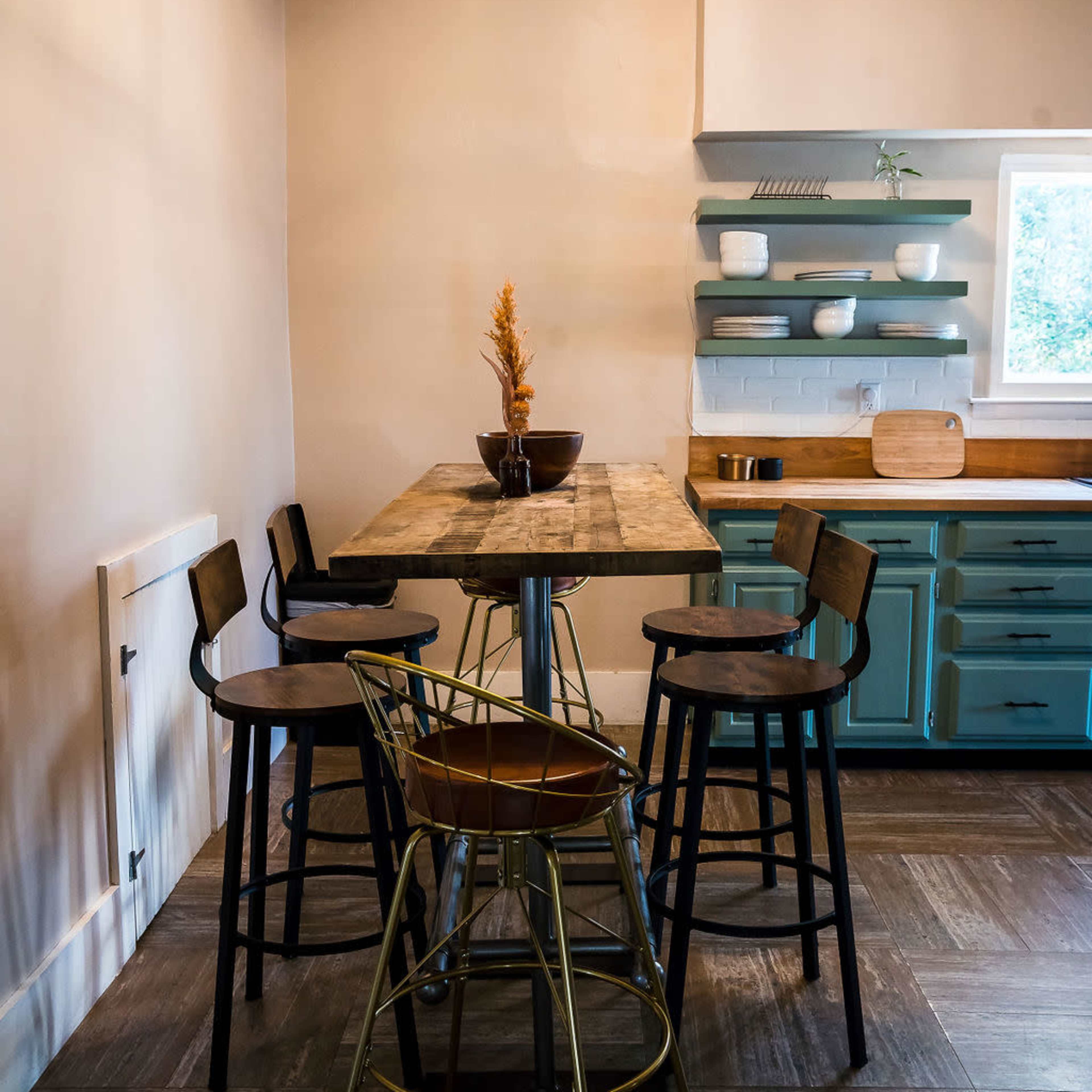 A small kitchen features a wooden table surrounded by four stools and a teal cabinetry setup.