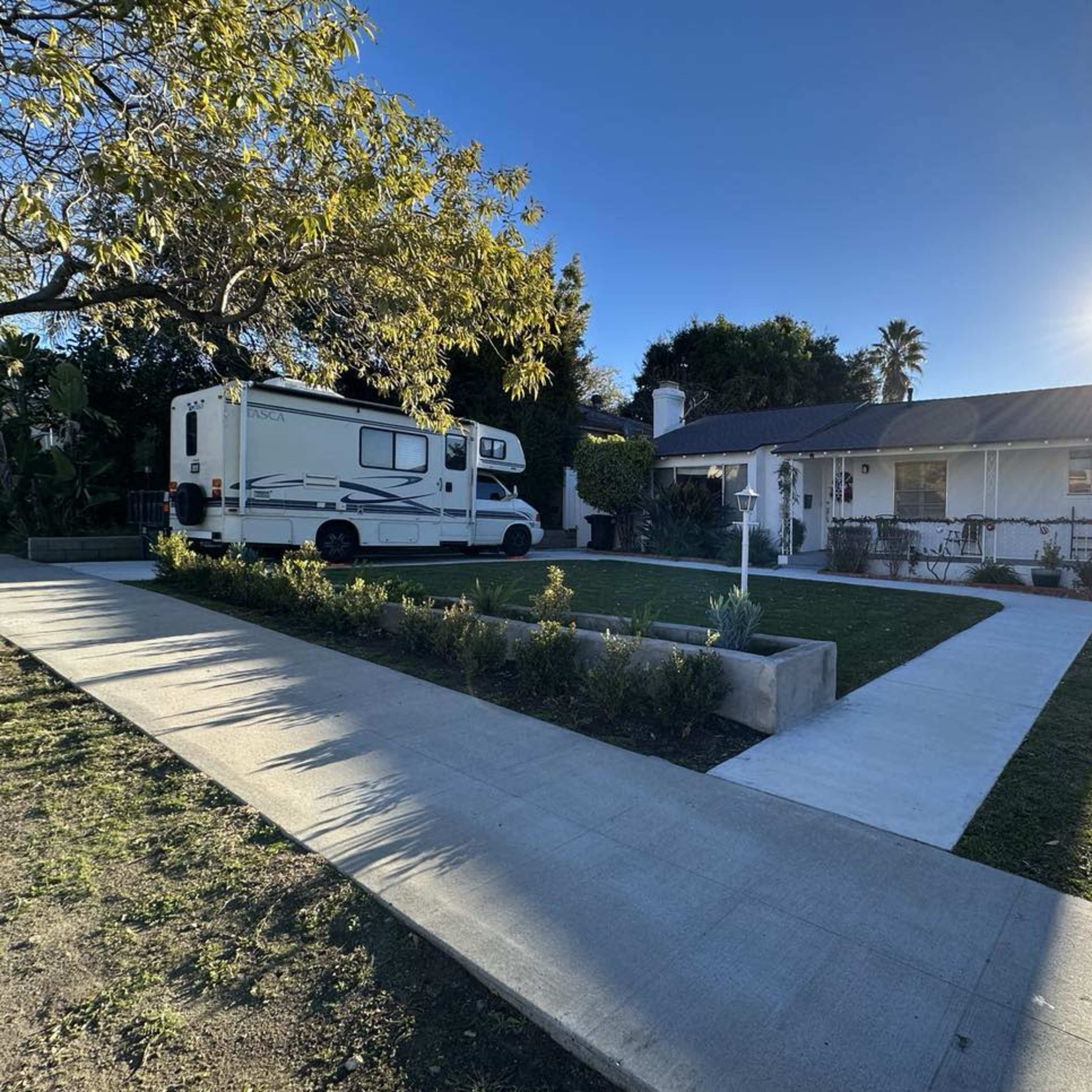 A recreational vehicle is parked in front of a single-story house with a landscaped yard and a clear blue sky overhead.