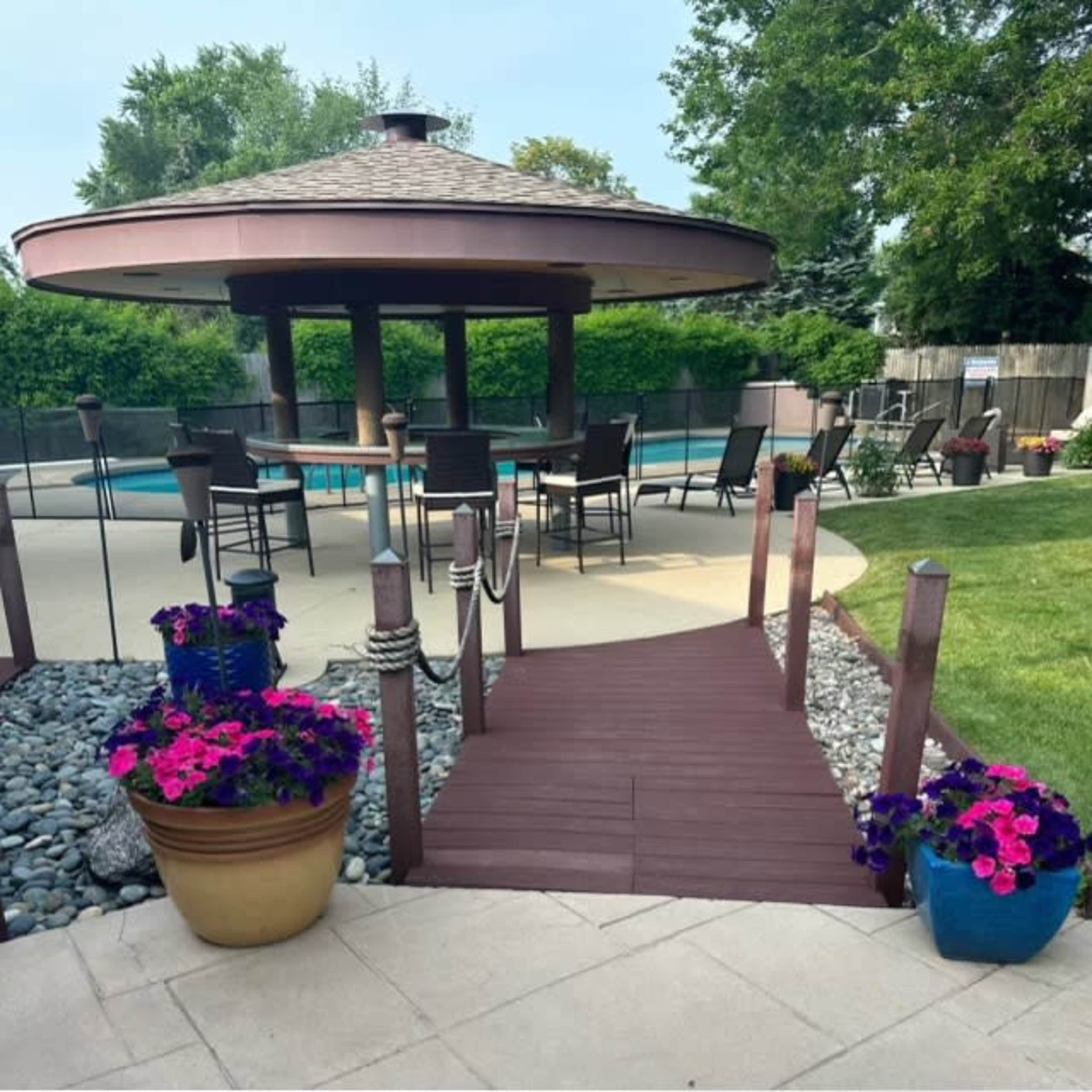 A wooden walkway leads to a round gazebo surrounded by seating and a pool in a landscaped backyard.