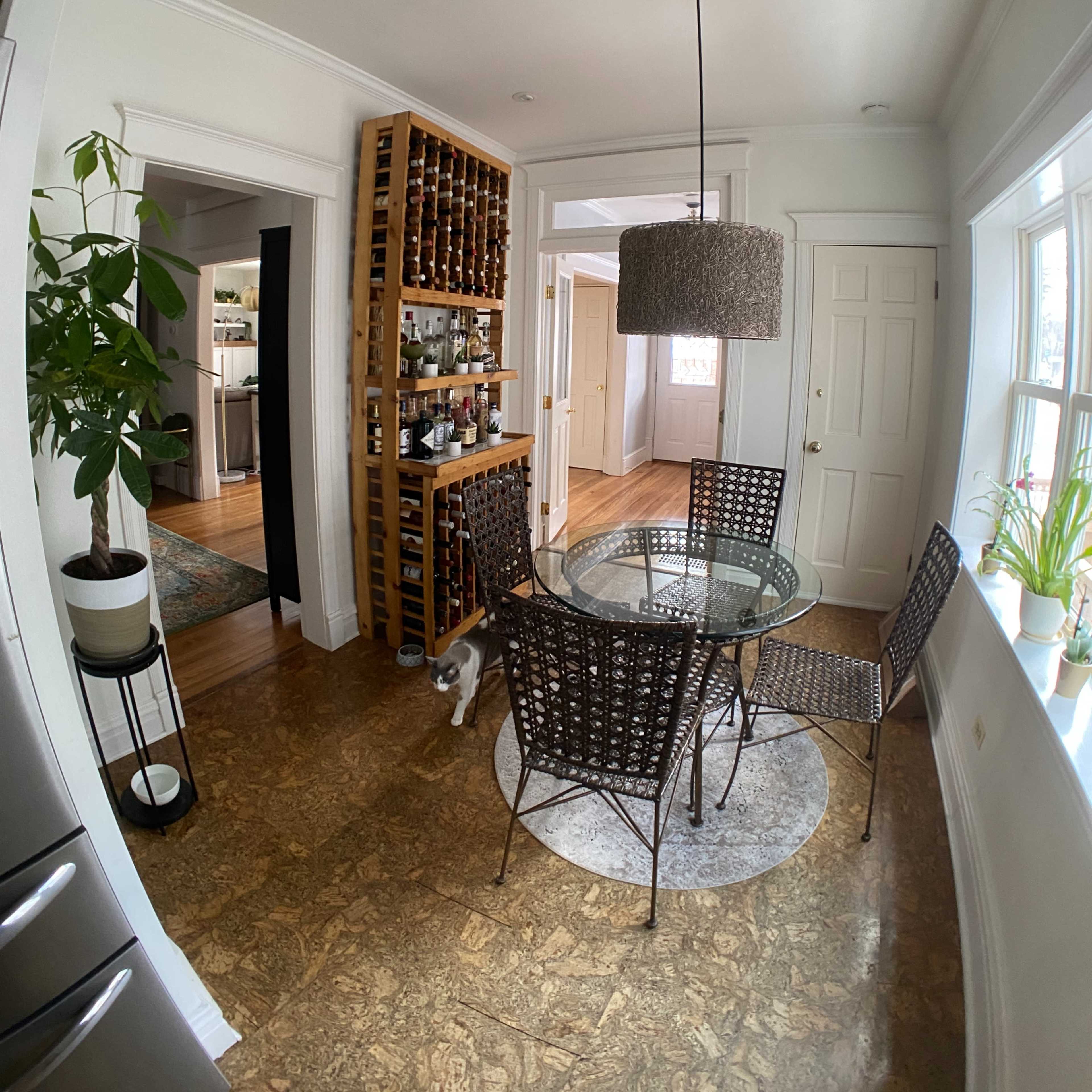 A small dining area features a round glass table surrounded by four wicker chairs, a wine rack filled with bottles, and a plant in the corner.