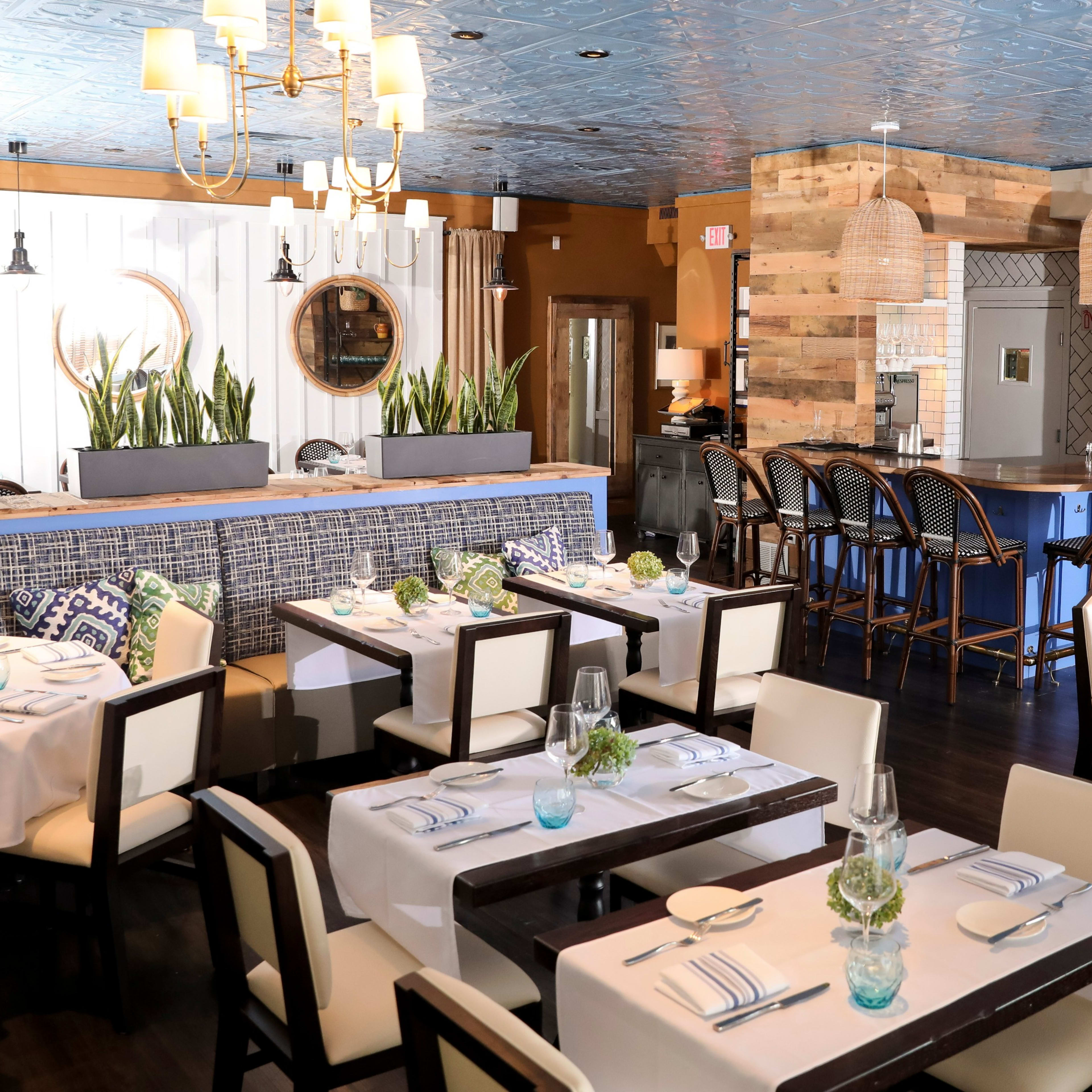 A modern restaurant interior with wooden tables, white tablecloths, and a blue ceiling, featuring a bar area and potted plants.