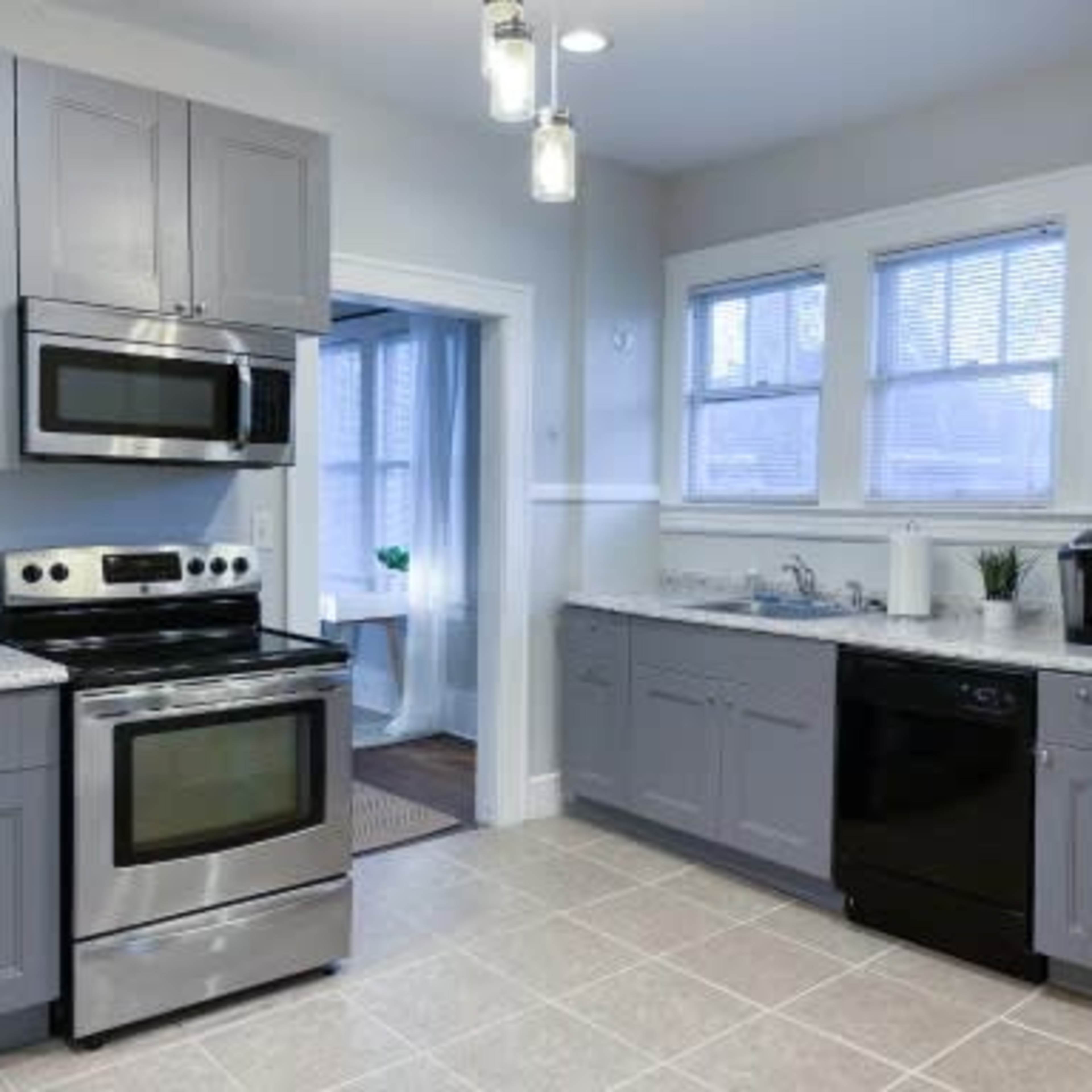 The kitchen features gray cabinets, stainless steel appliances, and tiled flooring with a view into a well-lit adjacent room.