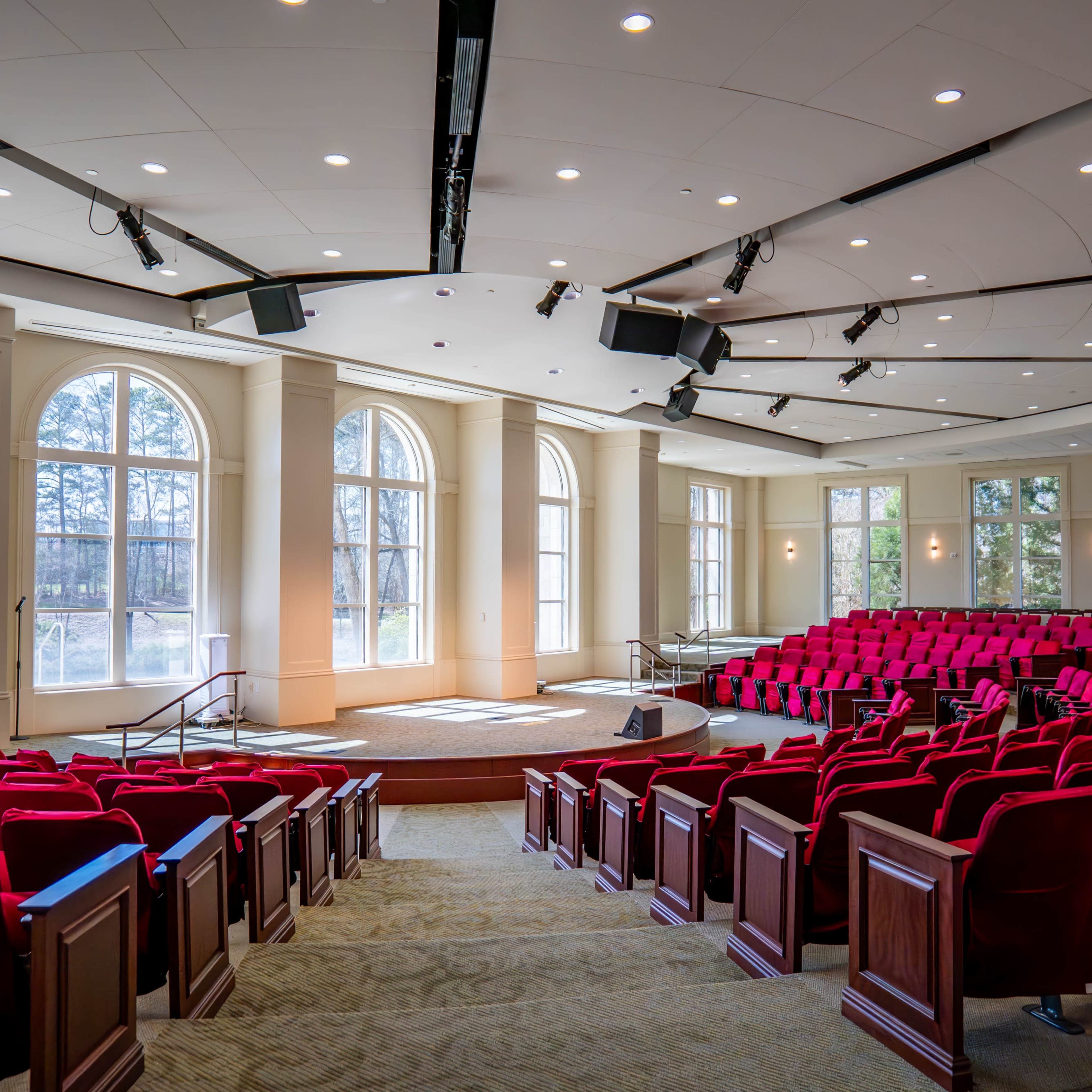 A spacious auditorium features rows of red seats facing a curved stage with large windows illuminating the space.