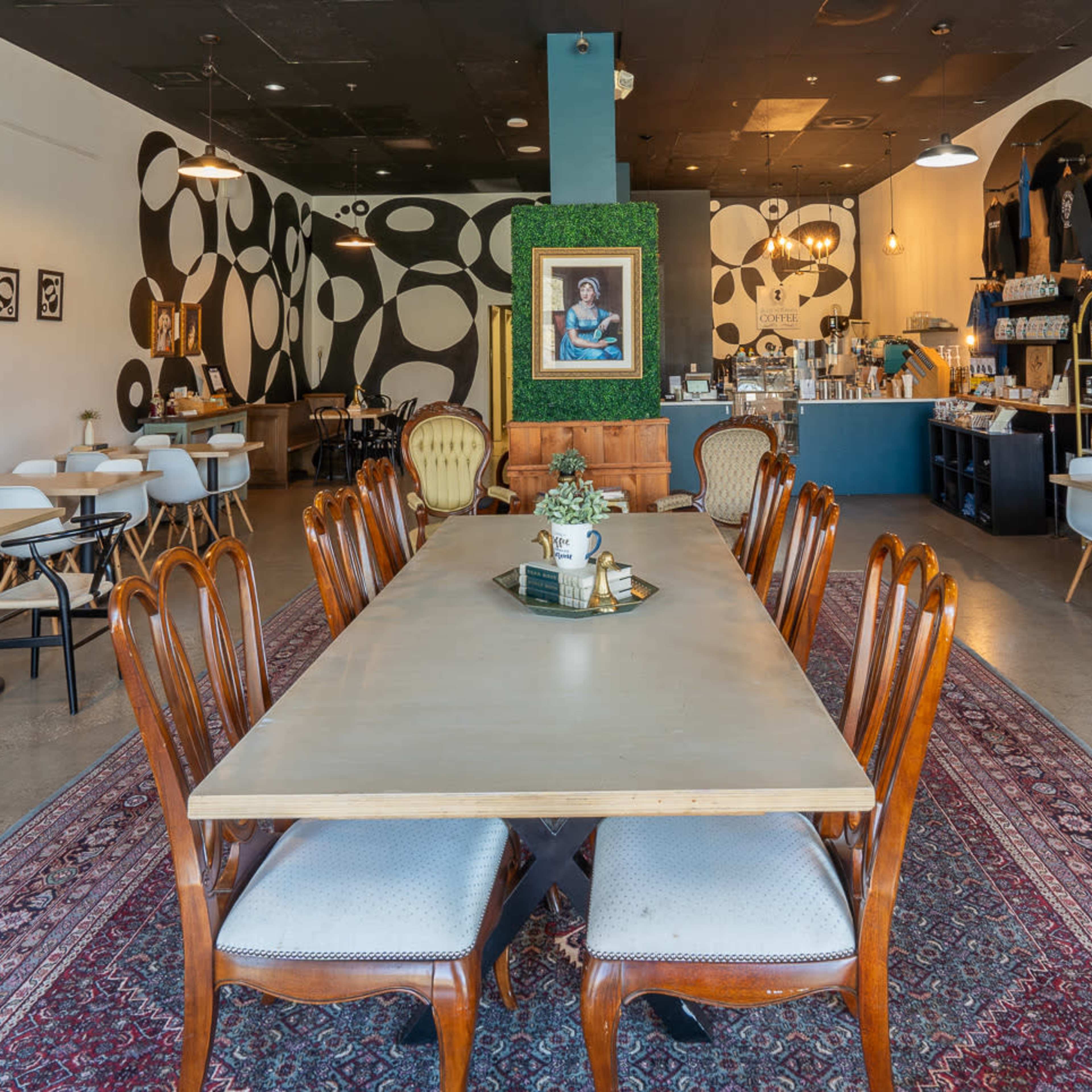 A contemporary café interior with a long wooden table, mixed seating arrangements, and patterned walls featuring abstract designs.