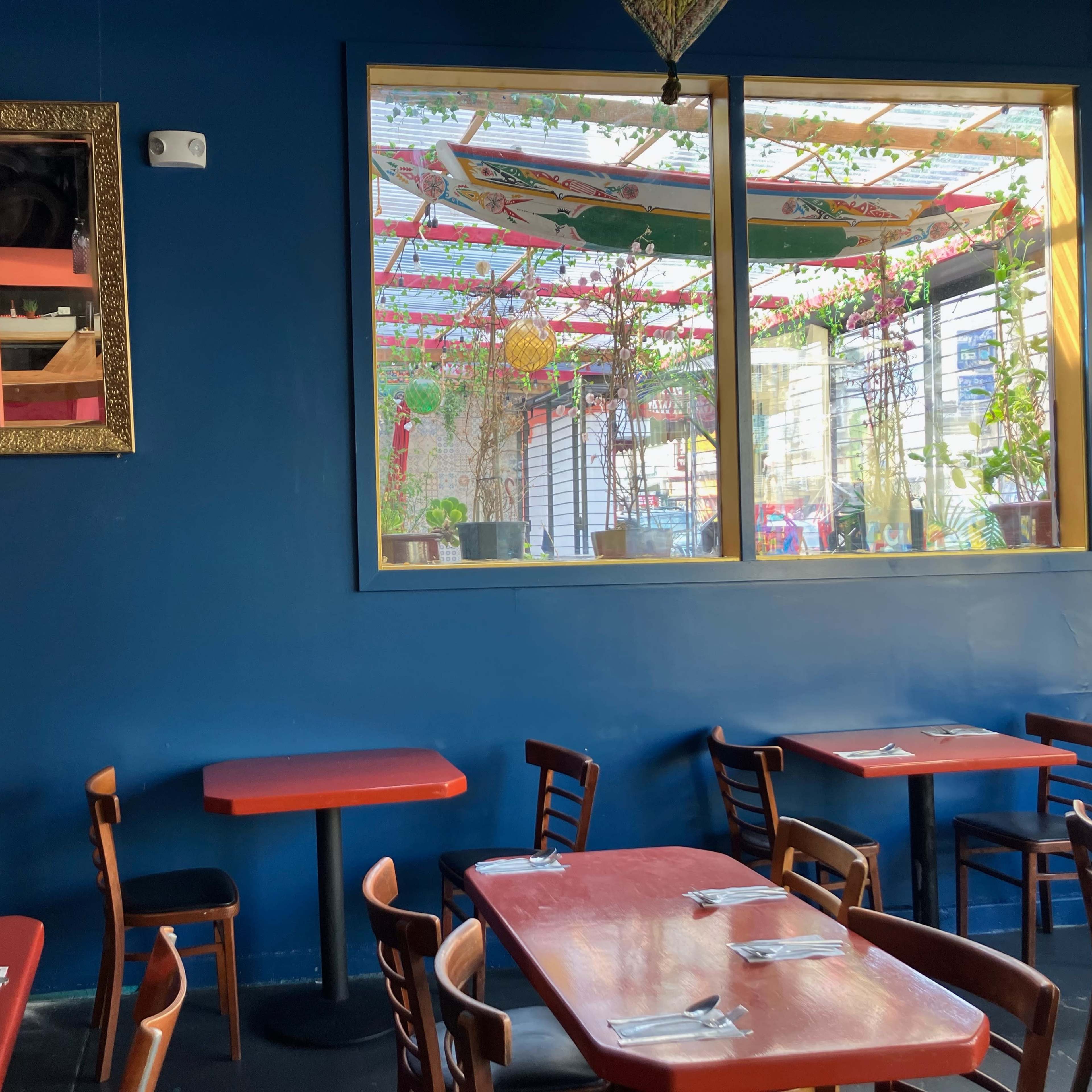 A restaurant interior with red tables and wooden chairs, featuring a large window that reveals a colorful outdoor area.