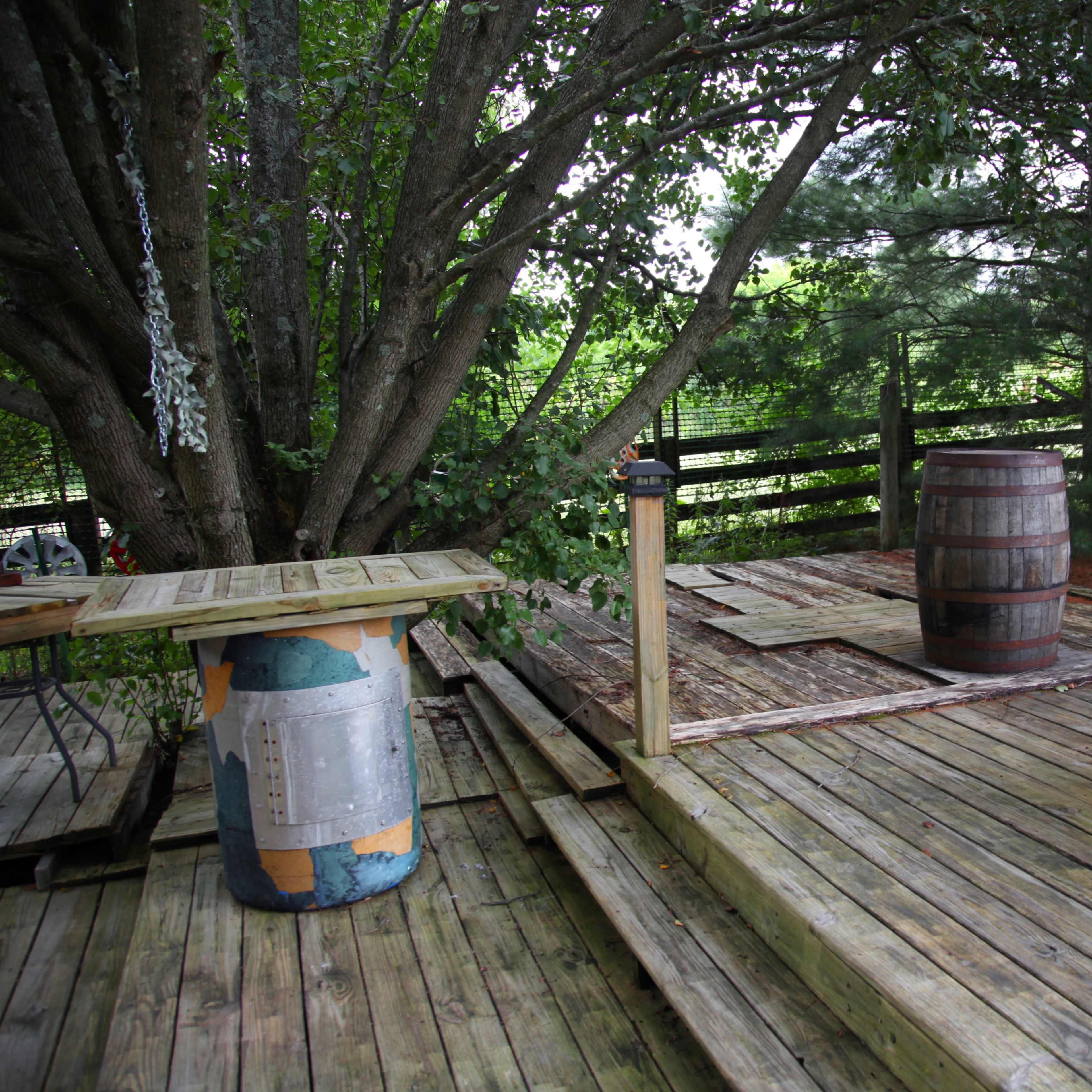 A wooden deck area features a barrel, a table made from an industrial spool, and surrounding greenery.