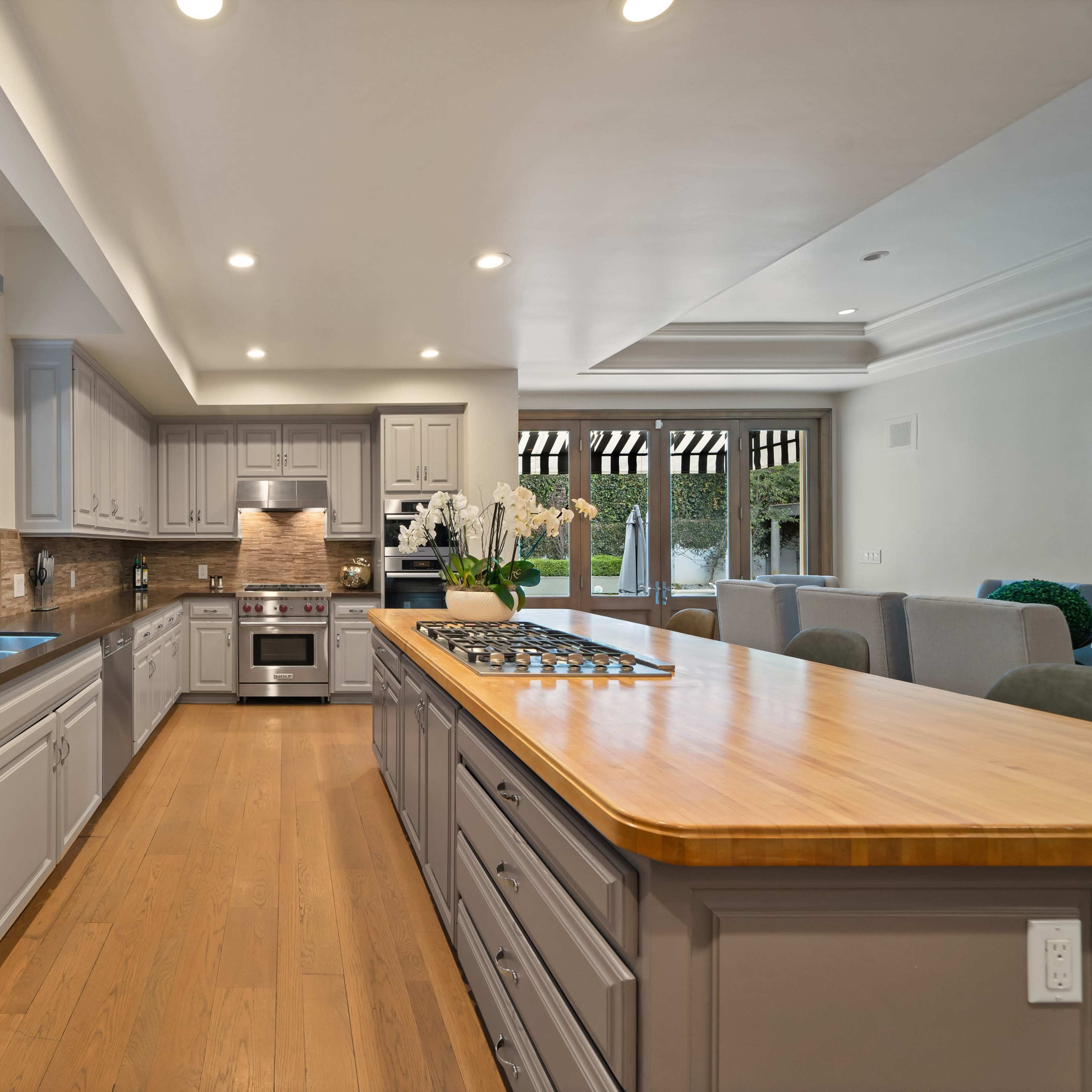A spacious kitchen features a central island with a wooden countertop, stainless steel appliances, and large windows overlooking a garden.