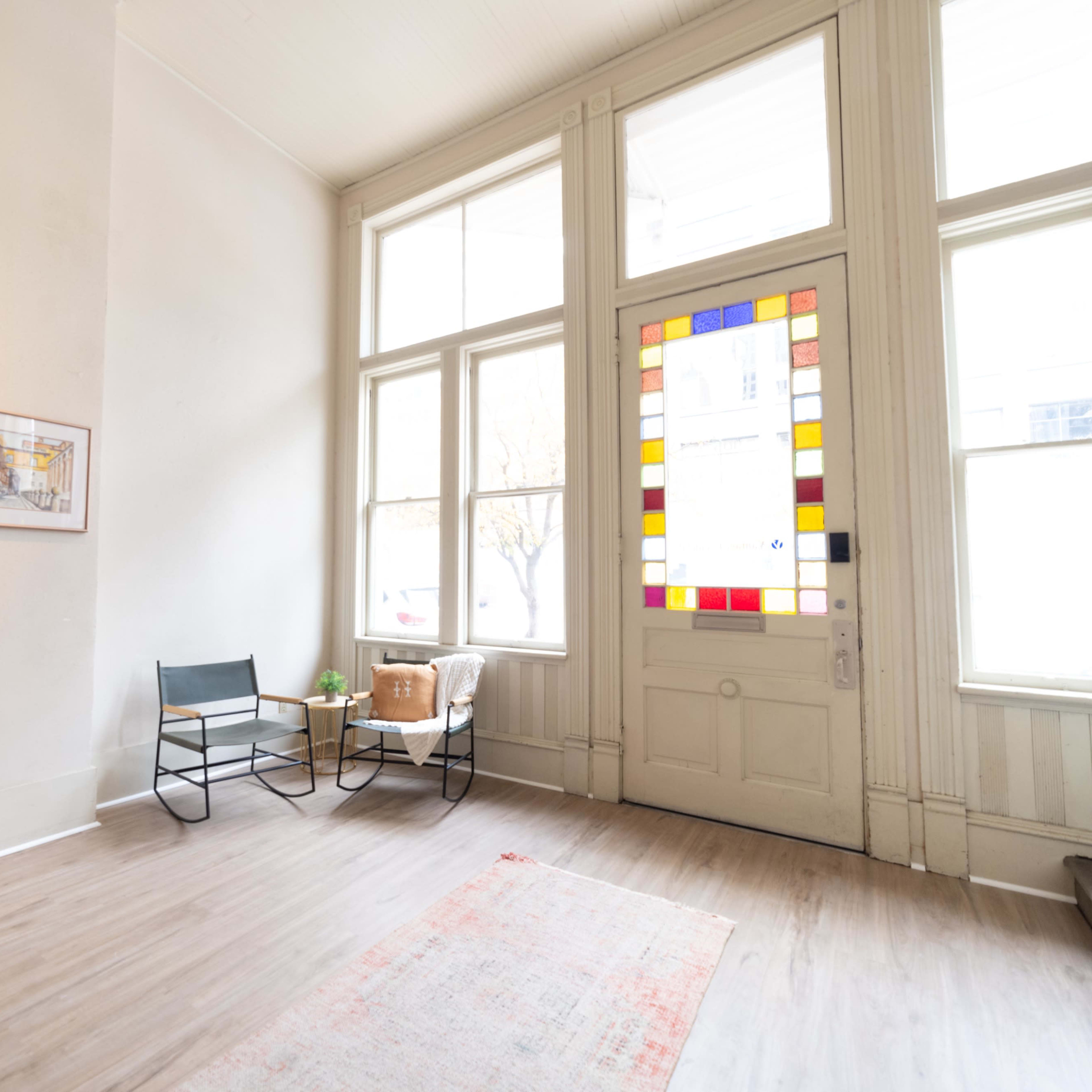 A bright foyer with large windows, a decorative door featuring colorful glass, a small seating area with a chair and a couch, a potted plant, and a light-colored wooden floor.
