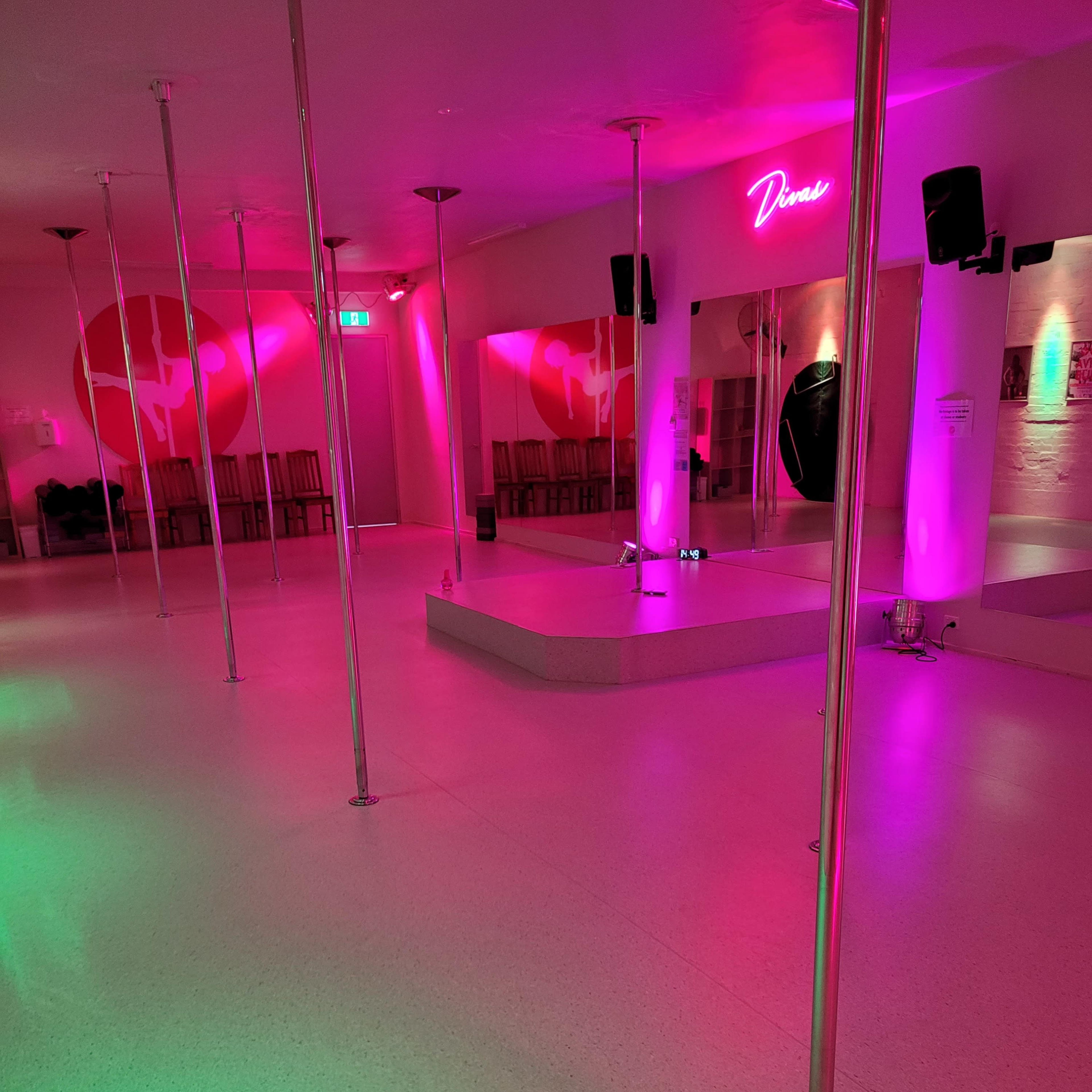 A dance studio with multiple vertical poles, illuminated by pink and green lighting, and a mirrored wall featuring the word "Dance."