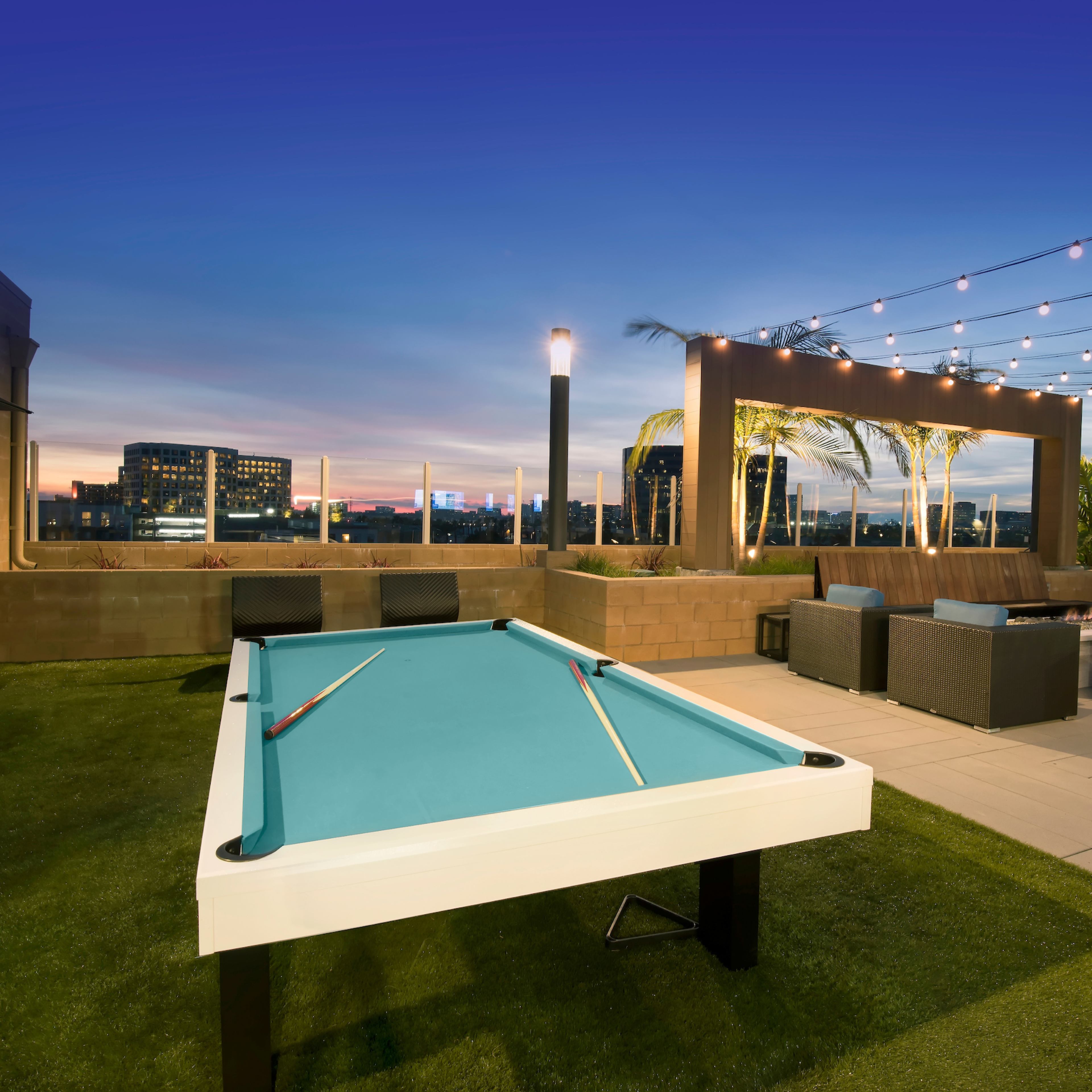 A rooftop terrace features a pool table surrounded by lounge seating and decorative string lights against a twilight sky.