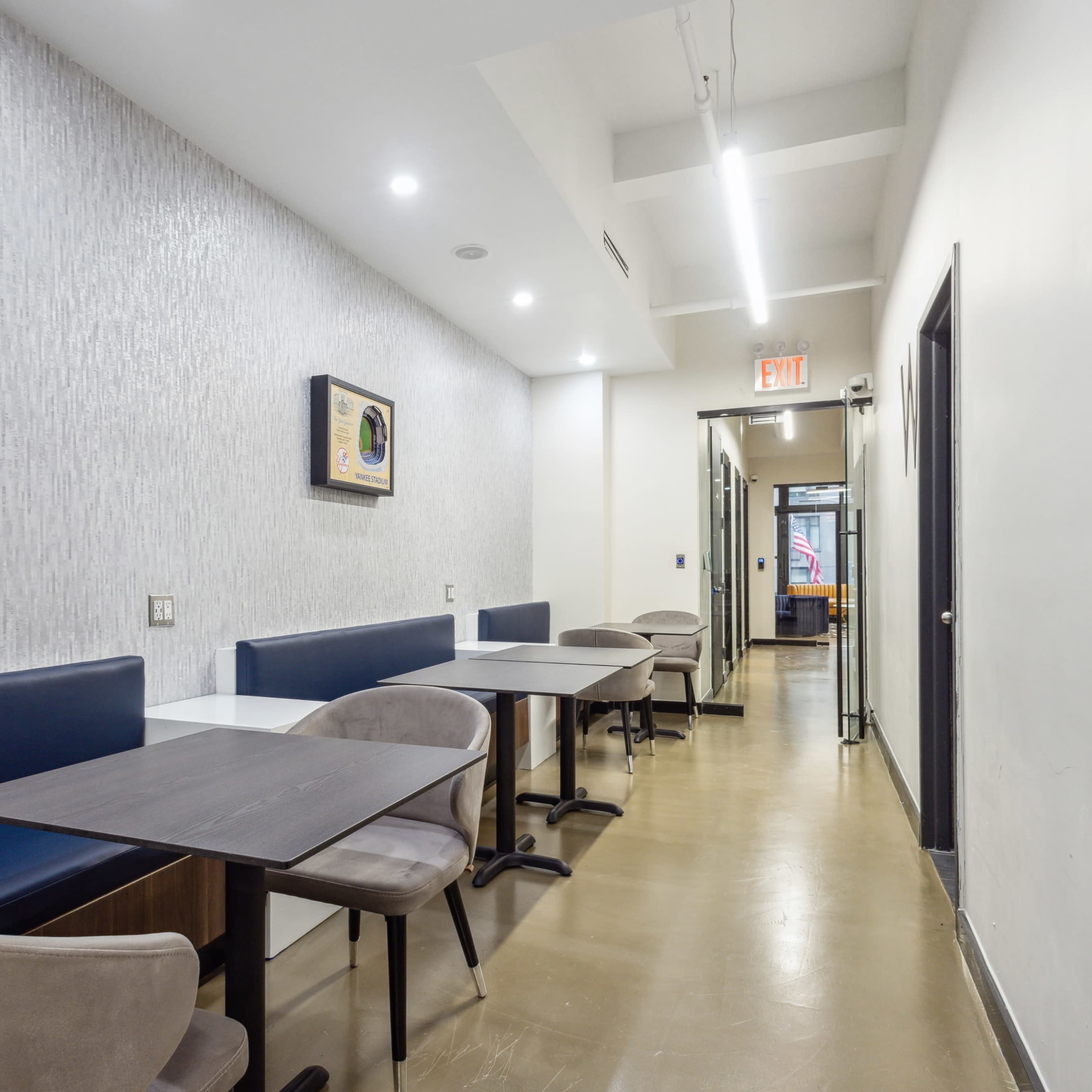 A narrow corridor features several tables and booths against the wall, illuminated by overhead lights, with doors leading to other rooms at the far end.