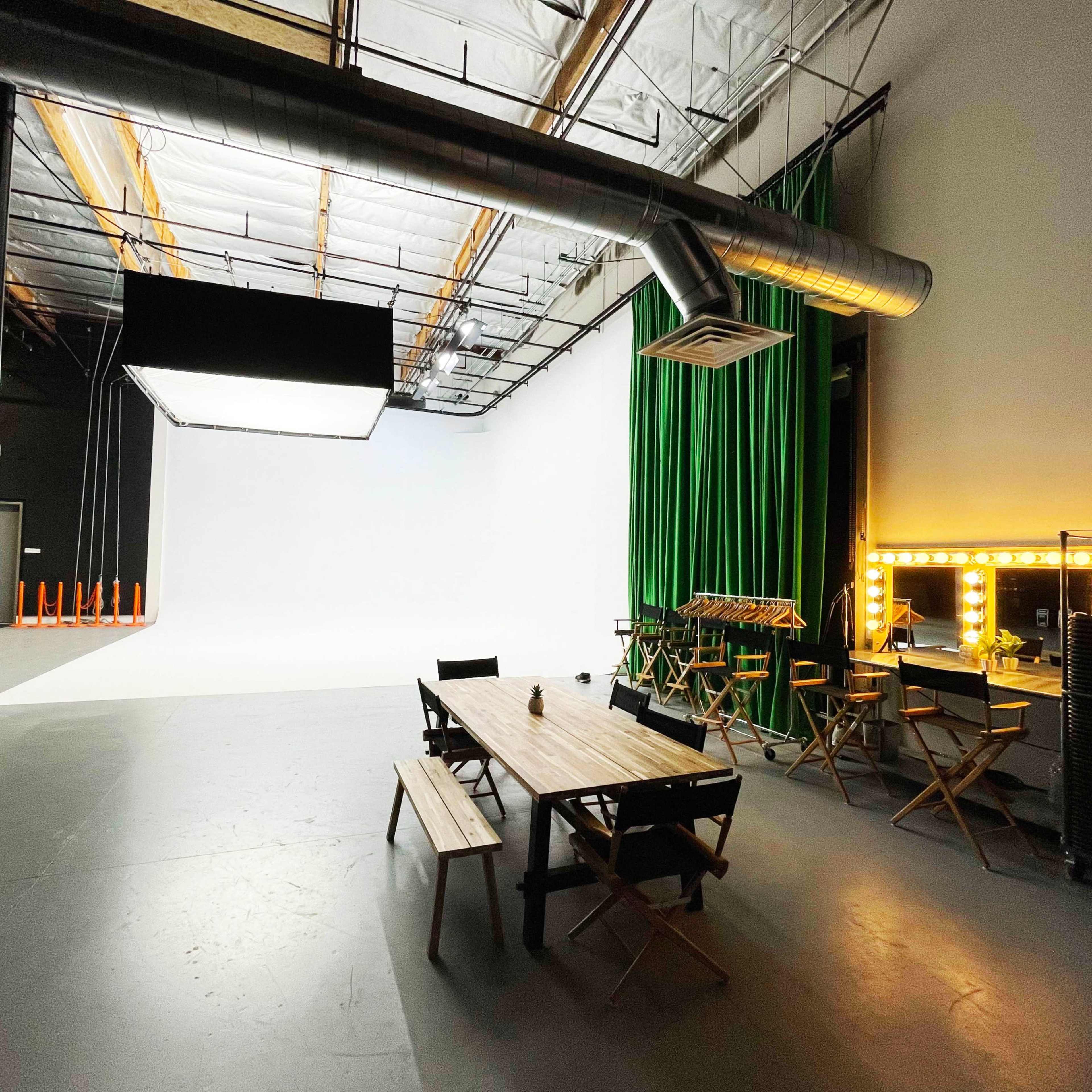 A spacious studio with a large white backdrop, green curtains, and various furniture including a wooden table and director's chairs.