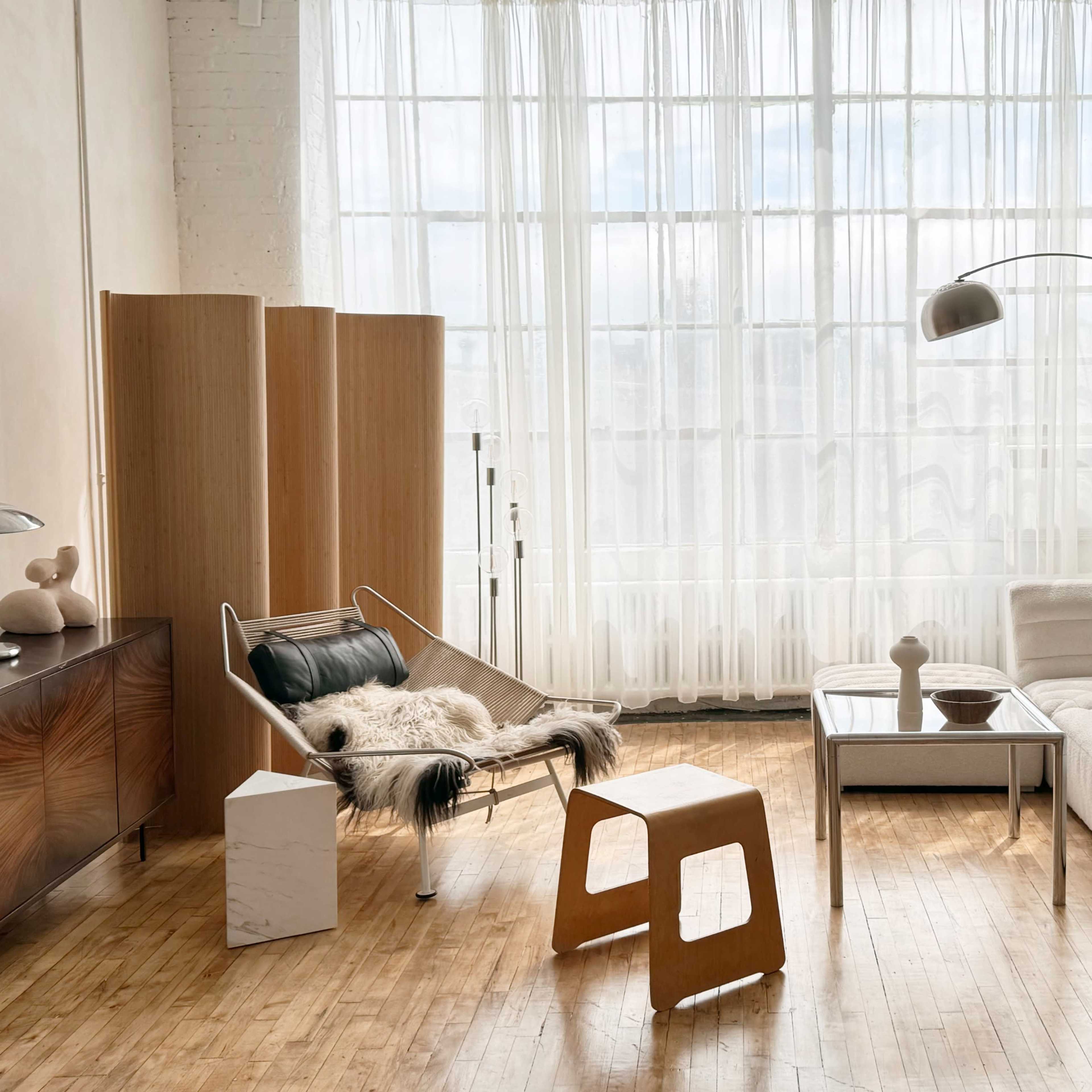 A modern living room with large windows, featuring a white sectional sofa, a sleek coffee table, and a wooden accent chair draped with a fur throw.