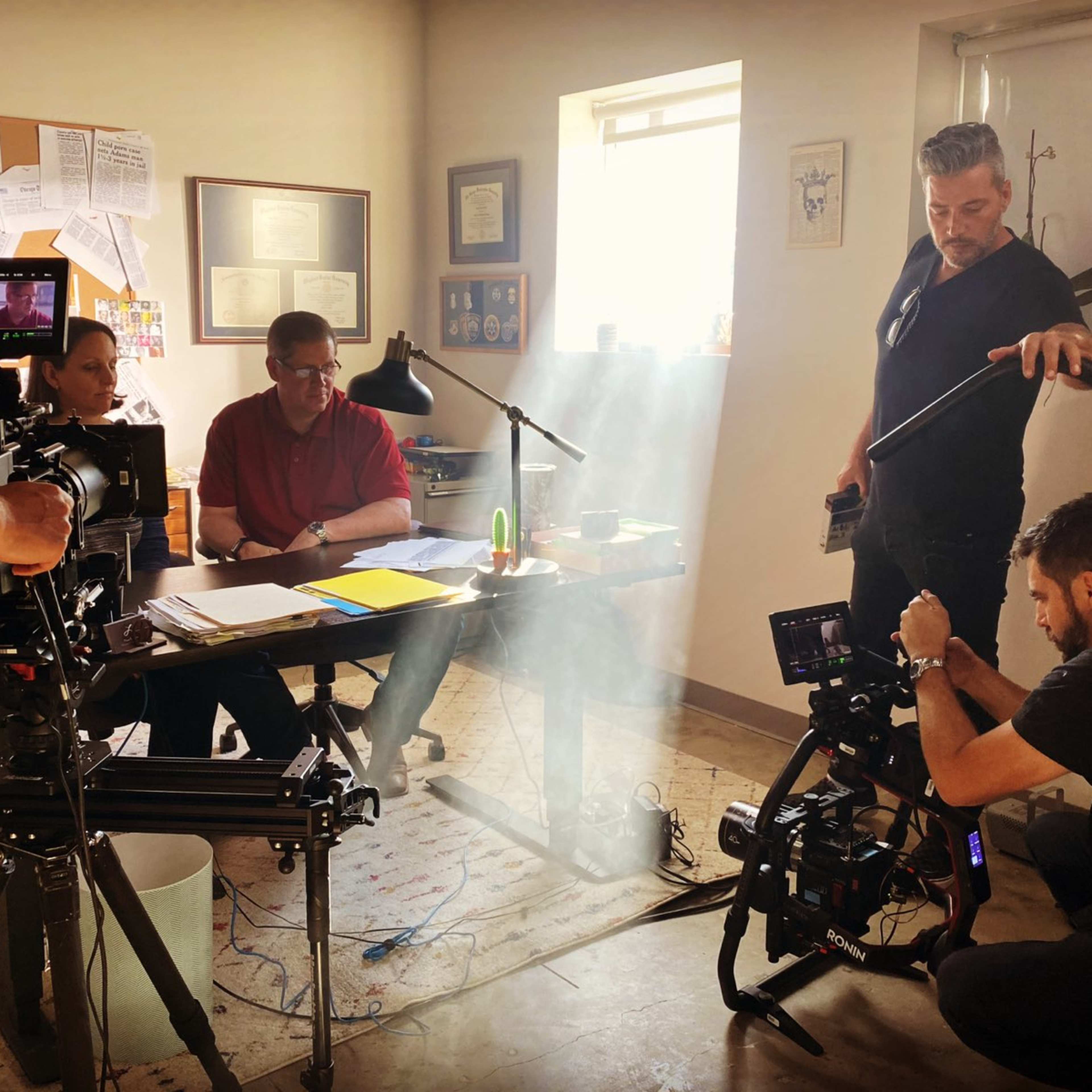 A film crew is capturing a scene in an office with a man sitting at a desk while another crew member adjusts equipment in a shaft of light.