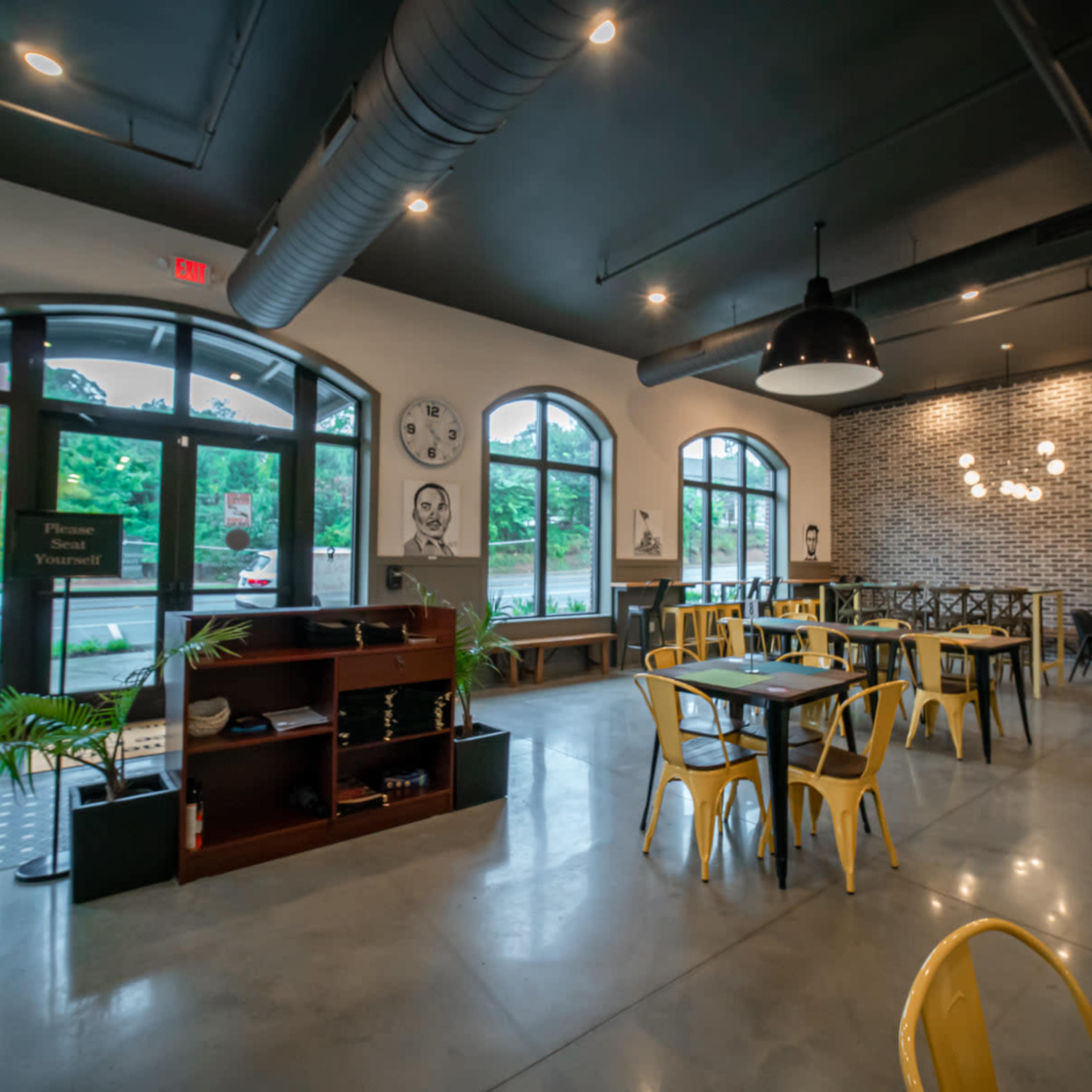 A modern café interior with industrial-style decor, featuring yellow chairs, wooden tables, and large windows allowing natural light to enter.