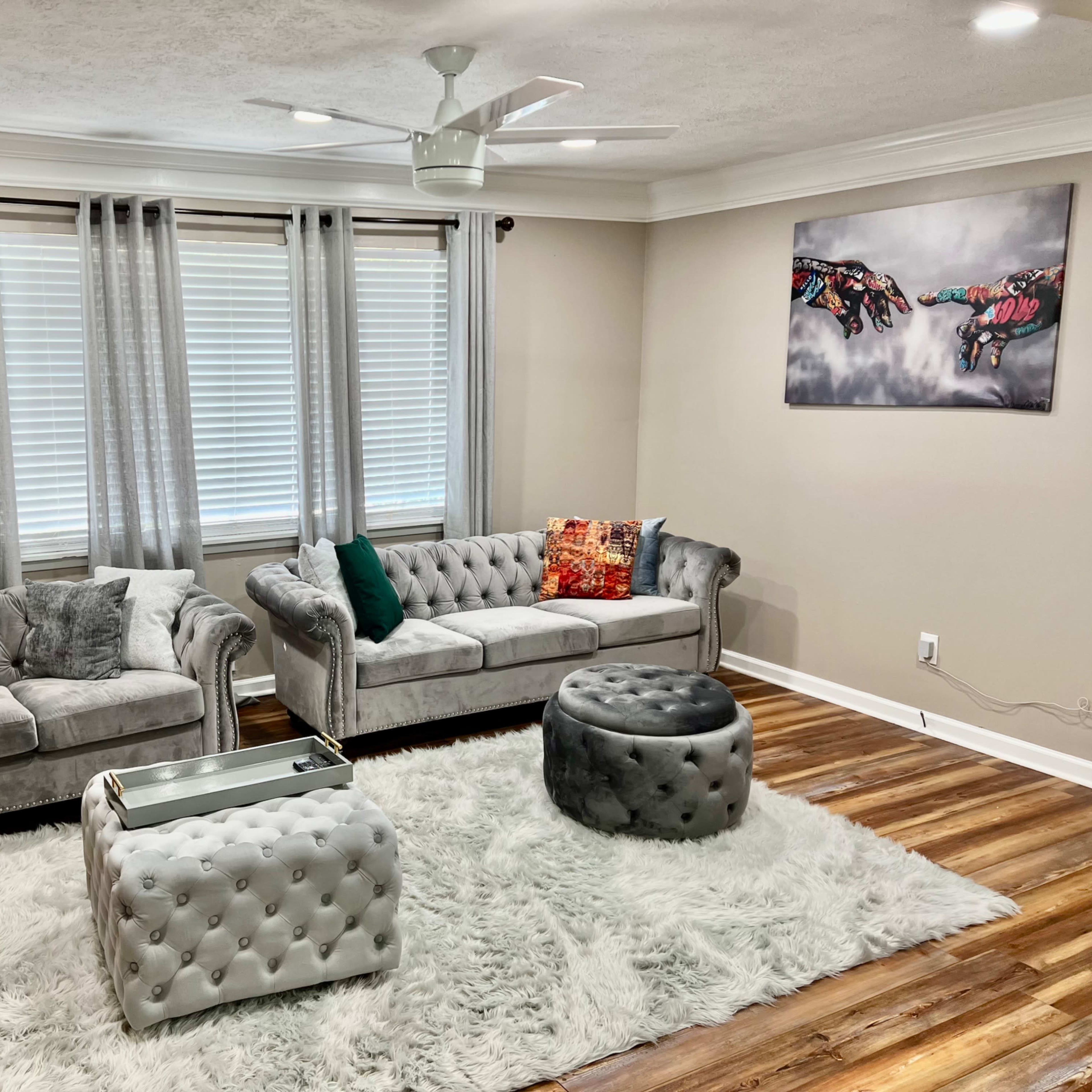 A bright living room features gray tufted sofas, a round ottoman, and a plush area rug set against light walls and large windows.