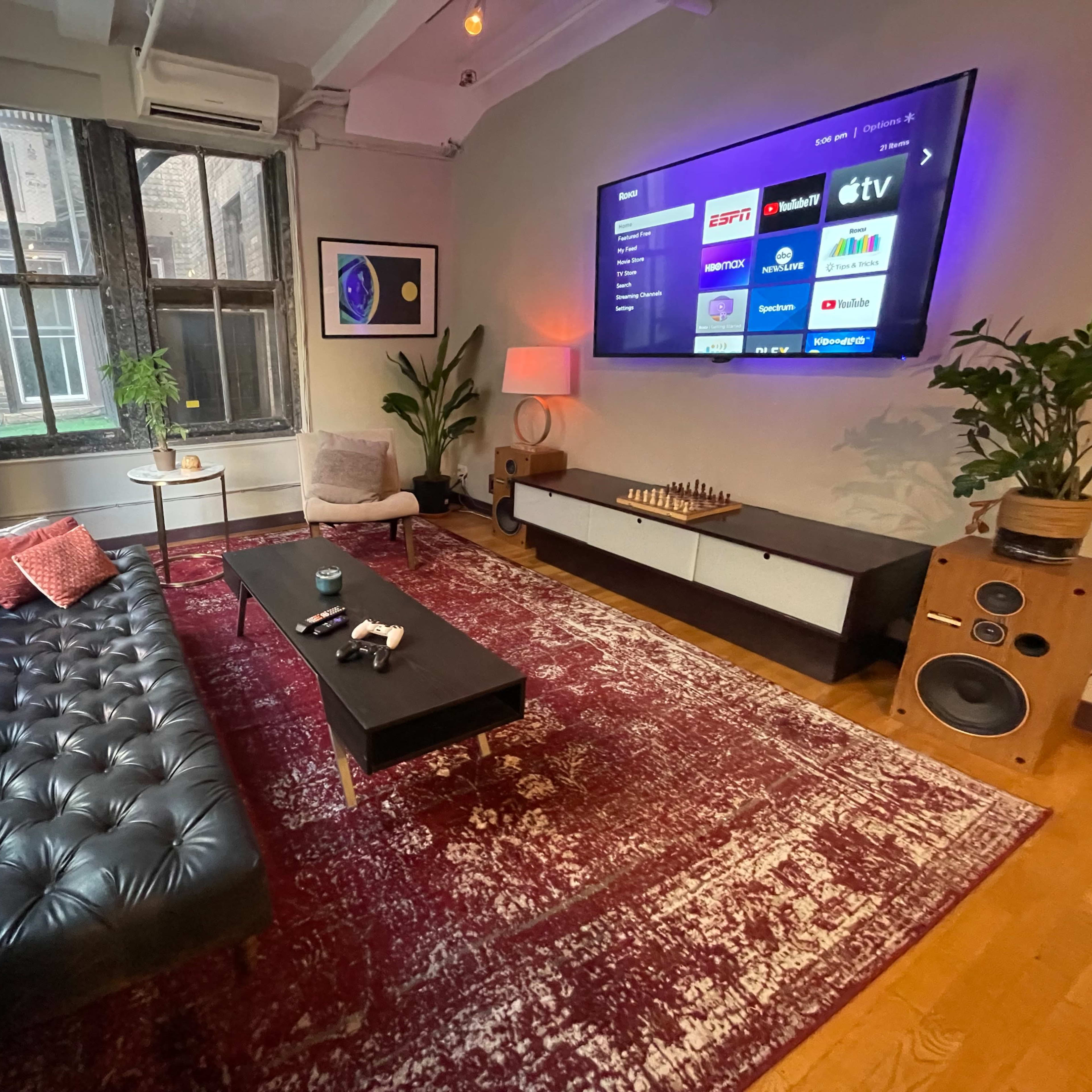 A cozy living room with a tufted black sofa, a coffee table, and a large TV mounted on the wall, surrounded by natural light from two windows.