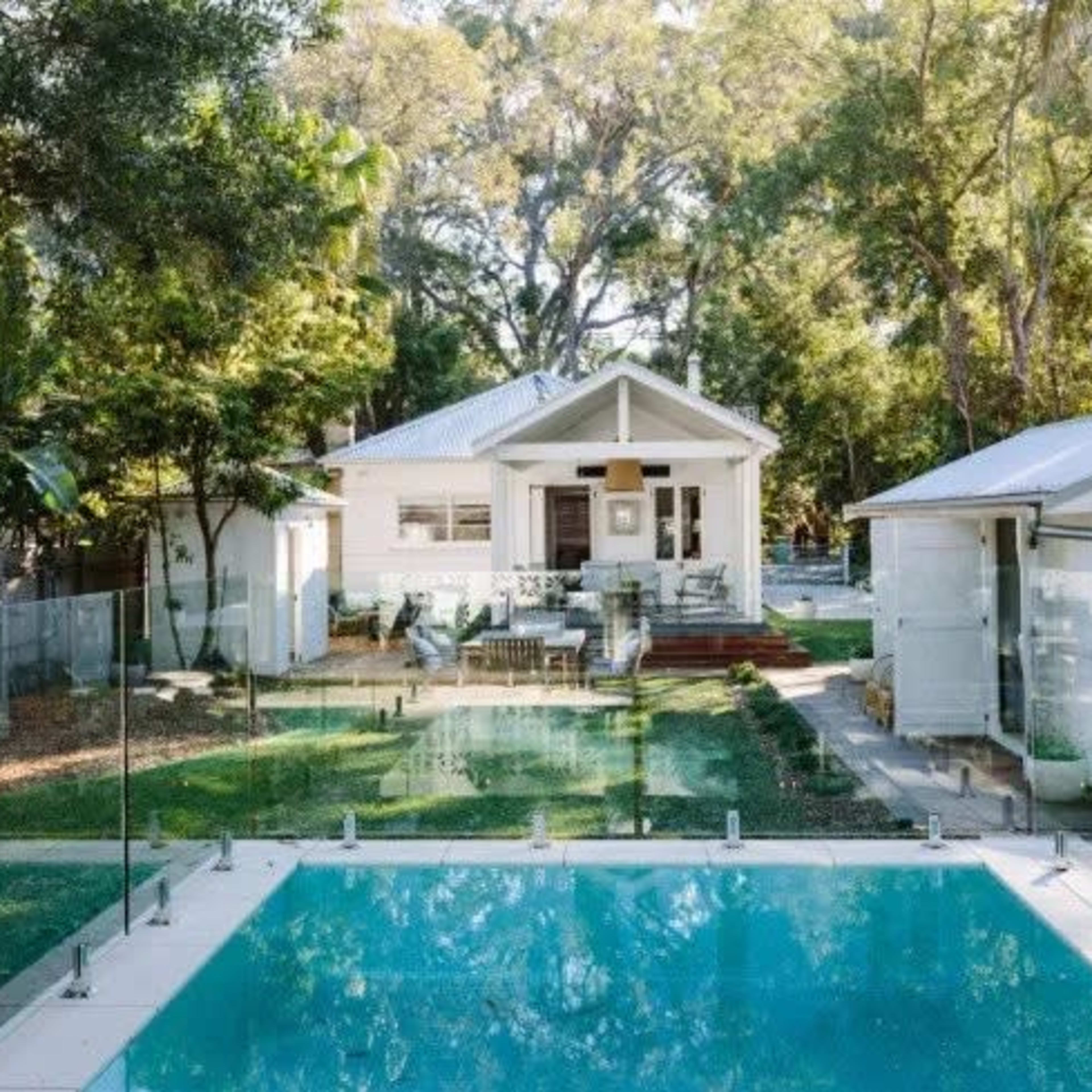 A clear swimming pool is situated in front of a modern house surrounded by greenery and a glass fence.