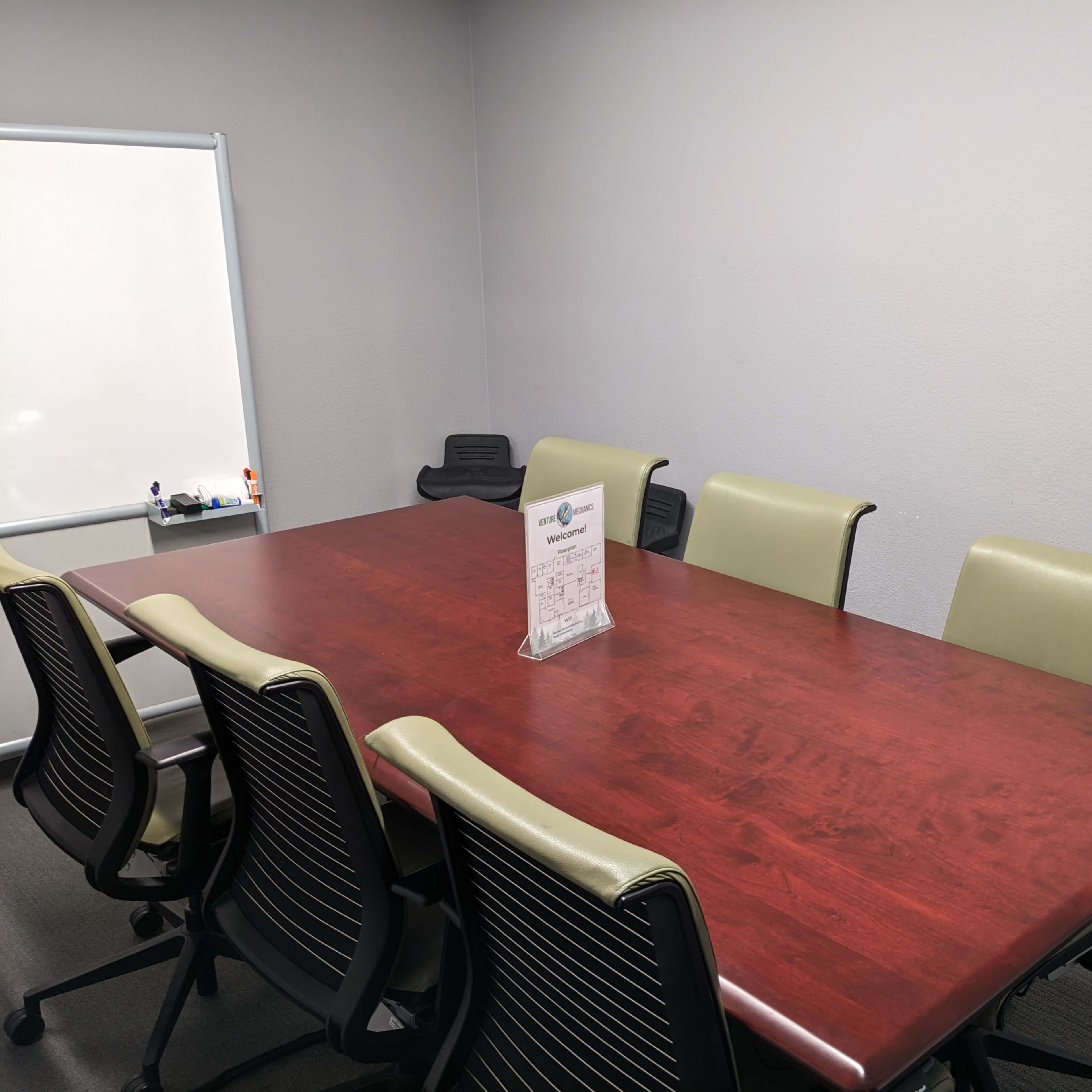 A rectangular wooden conference table surrounded by six ergonomic chairs is set against a plain gray wall in a meeting room.