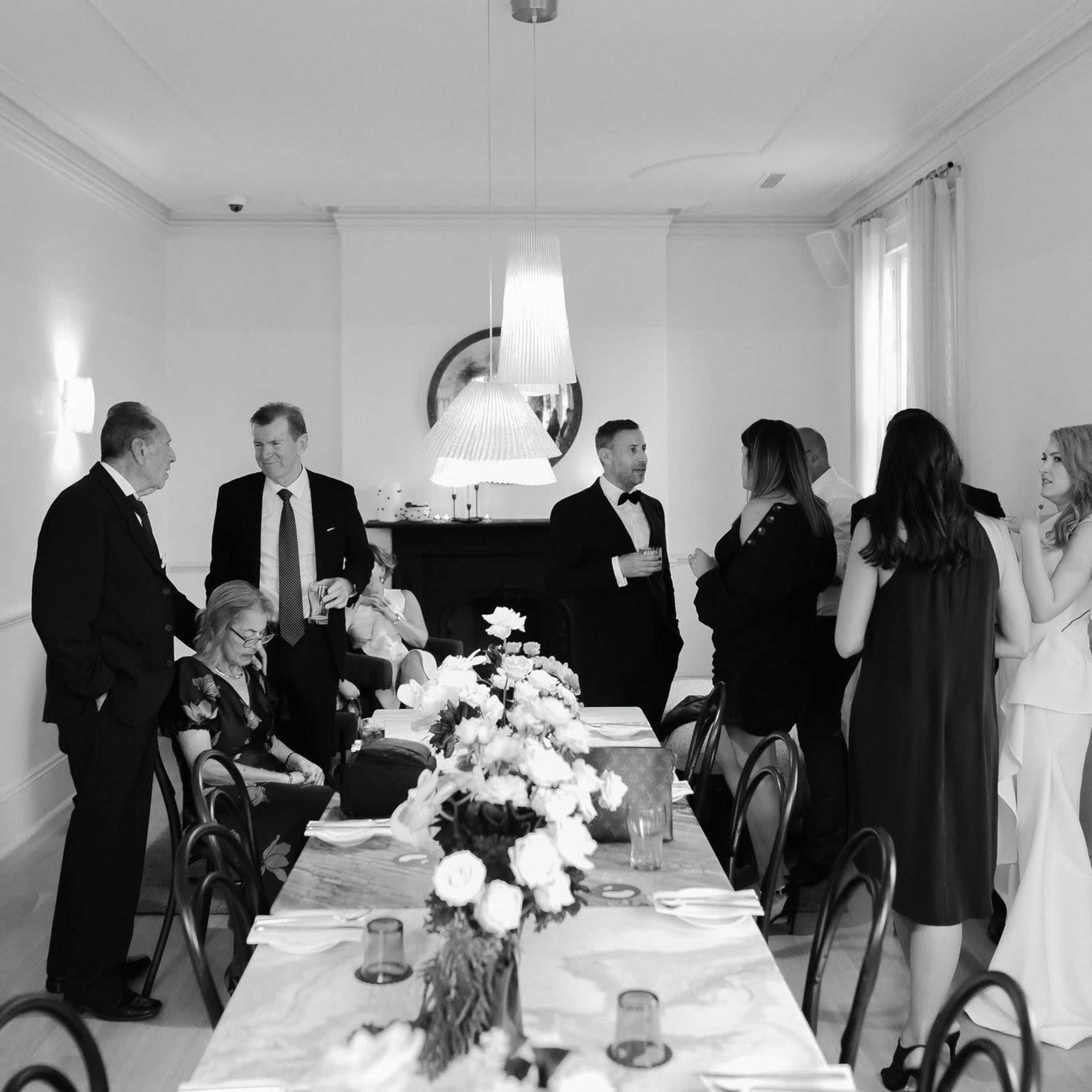 A group of elegantly dressed individuals socializes around a dining table adorned with flowers in a well-lit room.