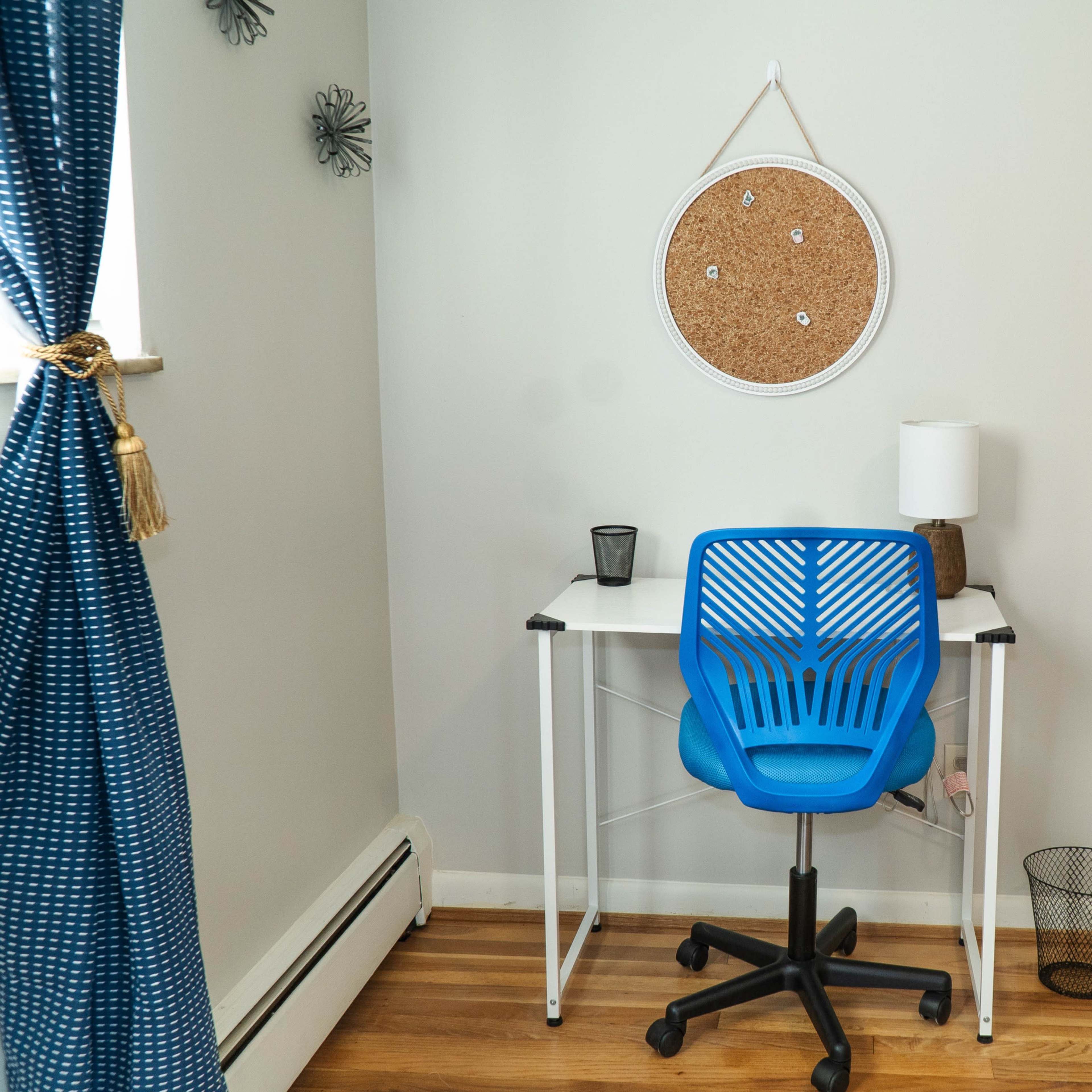 A blue office chair is positioned at a small white desk against a gray wall, with a cork board and lamp on the desk, and a closet visible in the background.