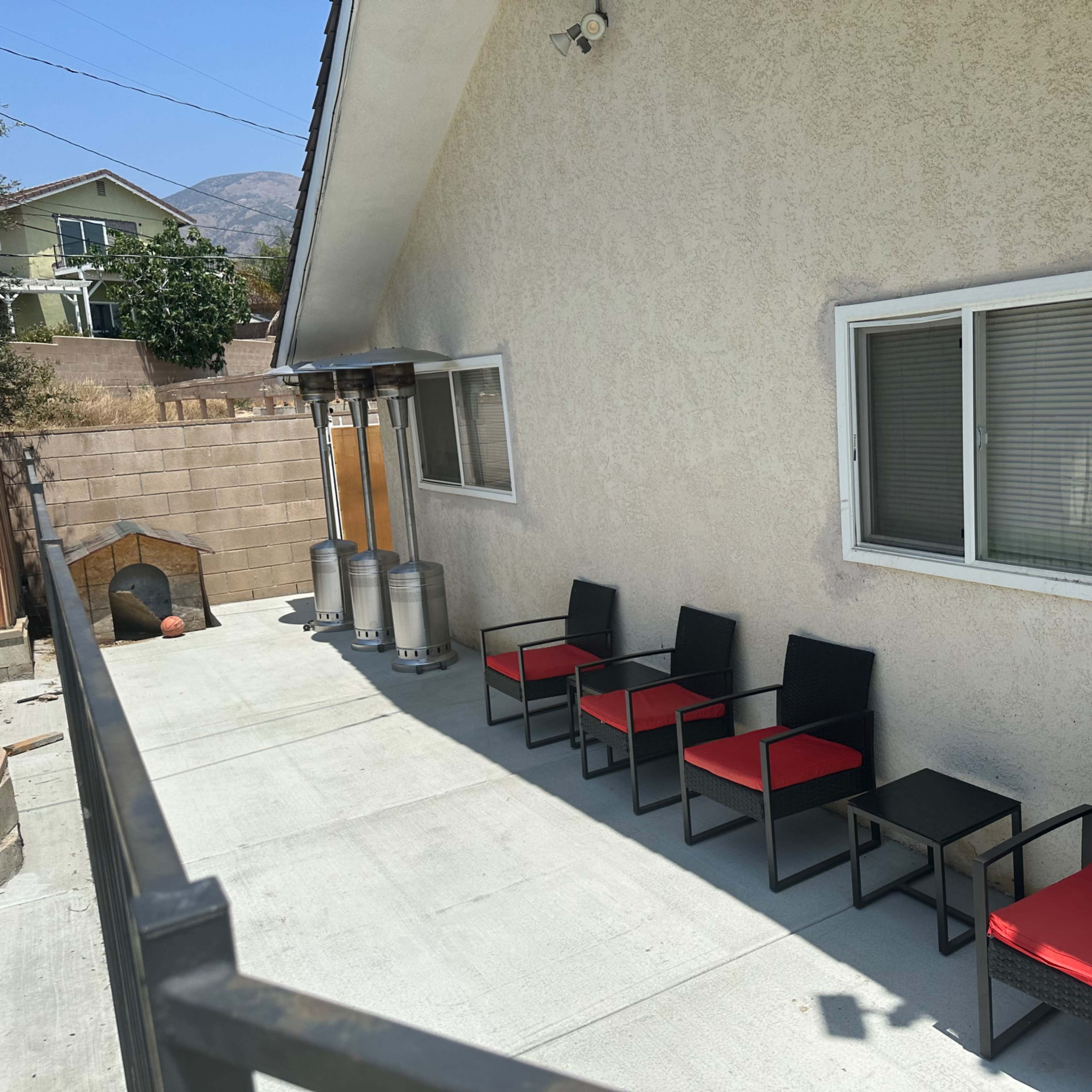A backyard patio with five black chairs featuring red cushions, a small table, and two patio heaters against a tan house wall.