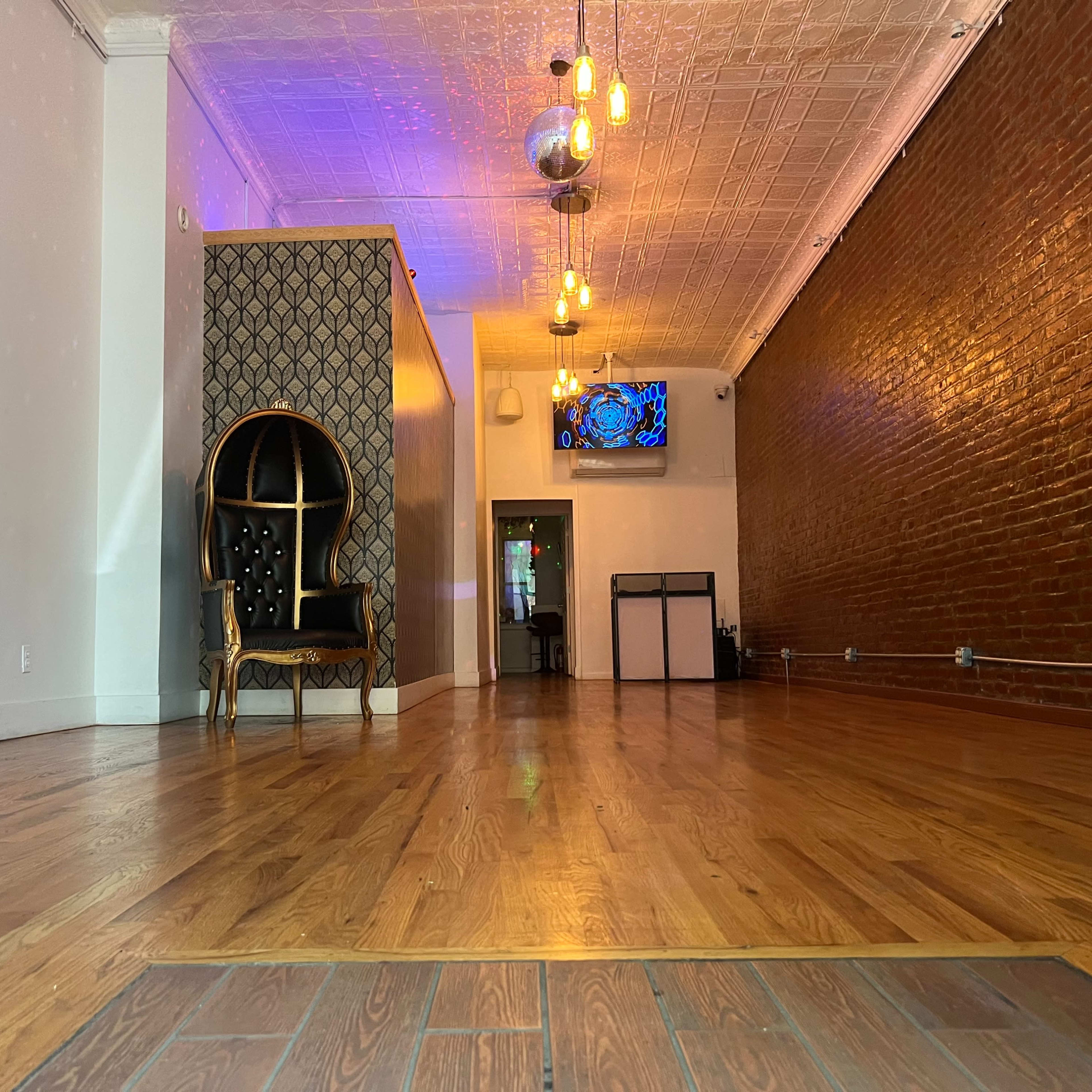 An empty interior space with wooden flooring, exposed brick walls, and a large decorative chair in the foreground.