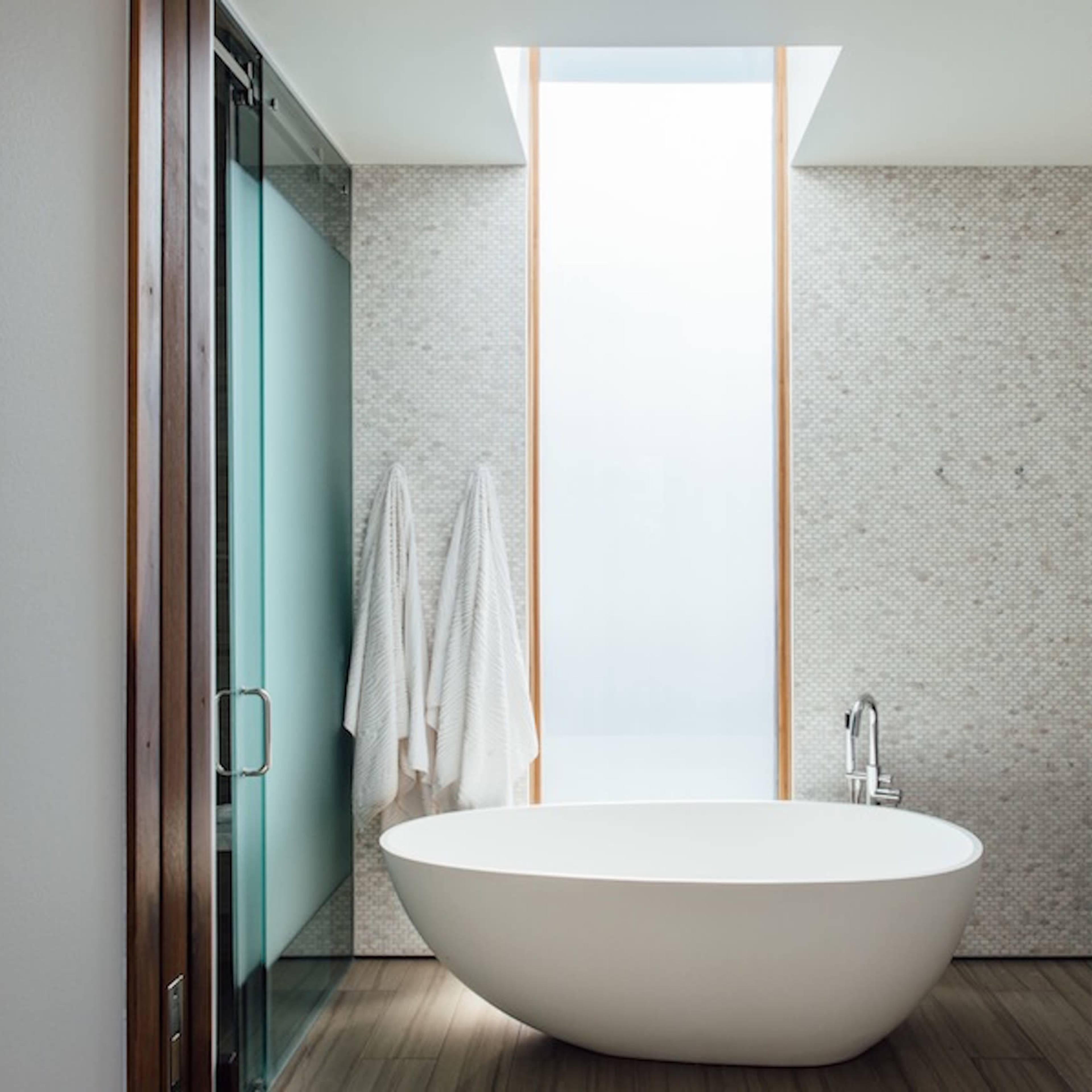 A modern bathroom features a freestanding oval bathtub, light stone walls, and a large window that allows natural light to enter.