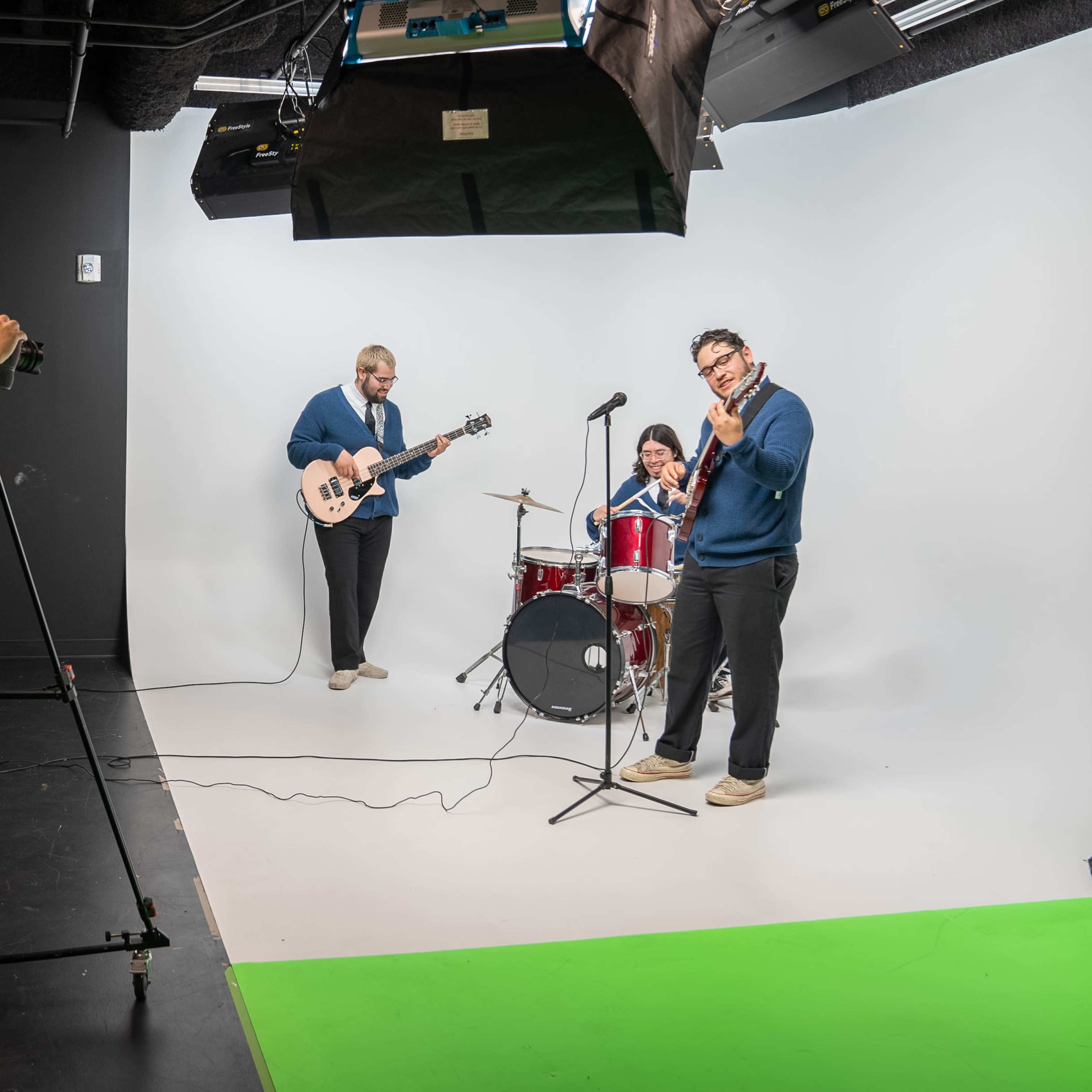 A band performs in a studio with a white backdrop while a camera operator records them.