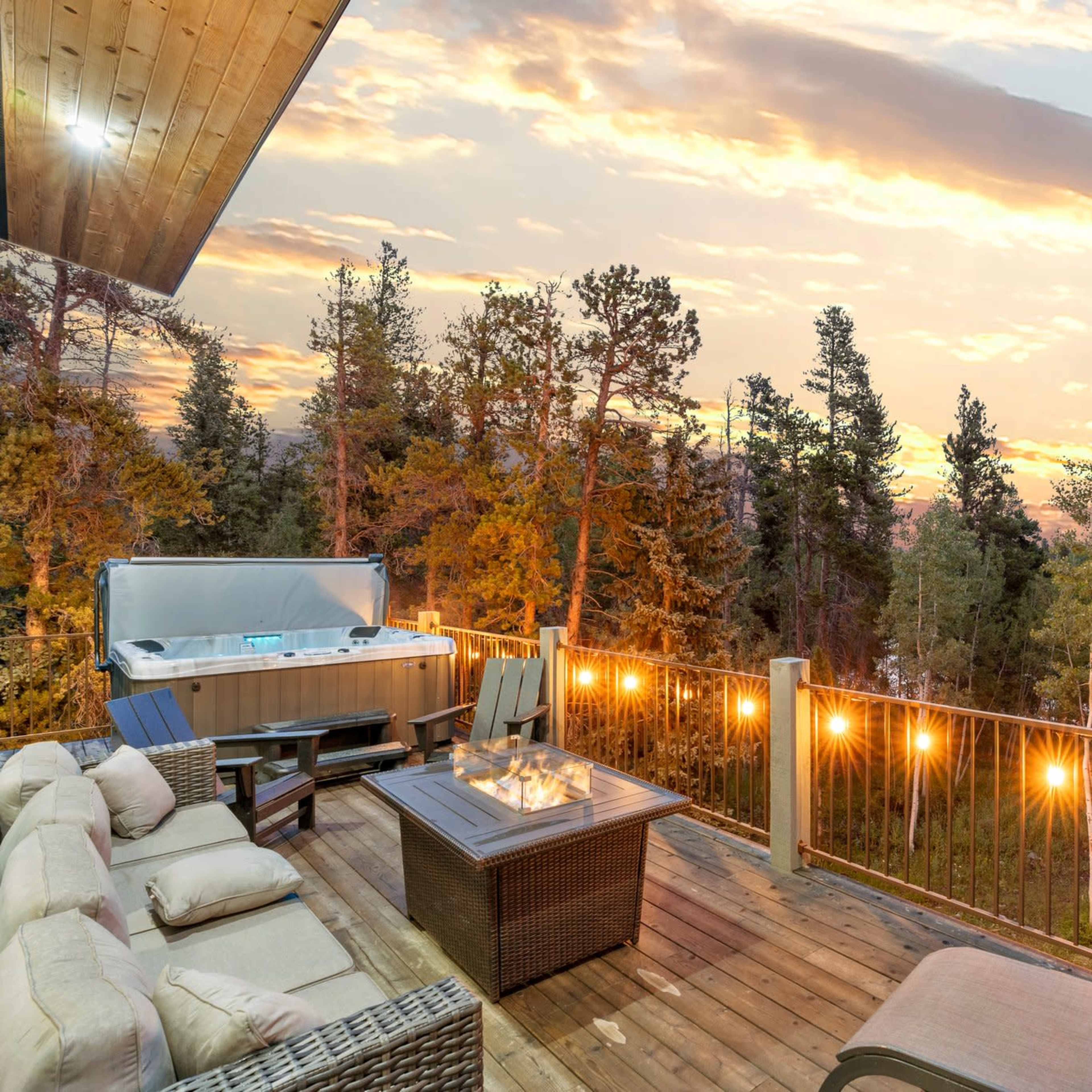 A wooden deck features a hot tub and seating area, surrounded by trees and overlooking a sunset sky.