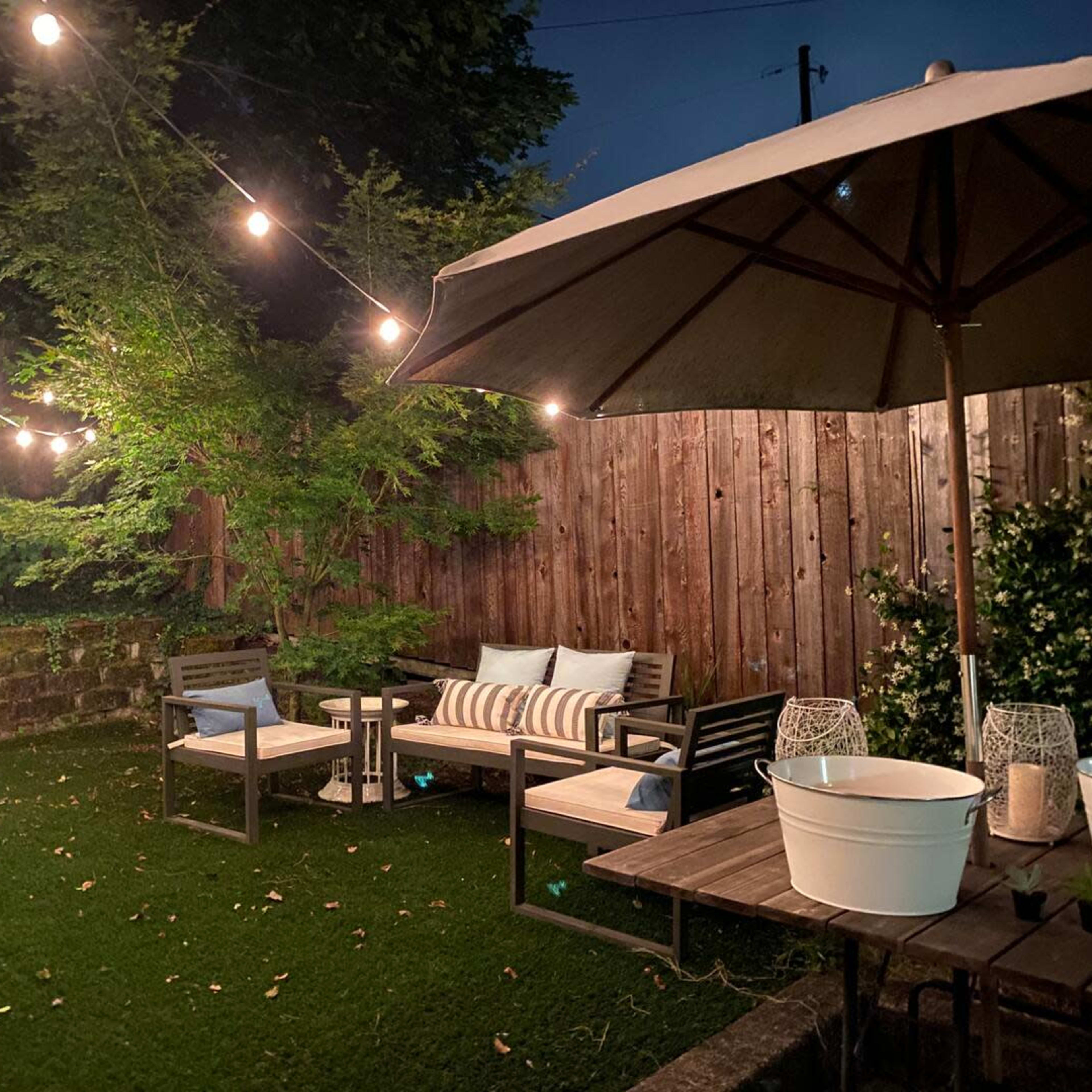 A cozy backyard scene features a seating area with two chairs and a table, surrounded by string lights and greenery.