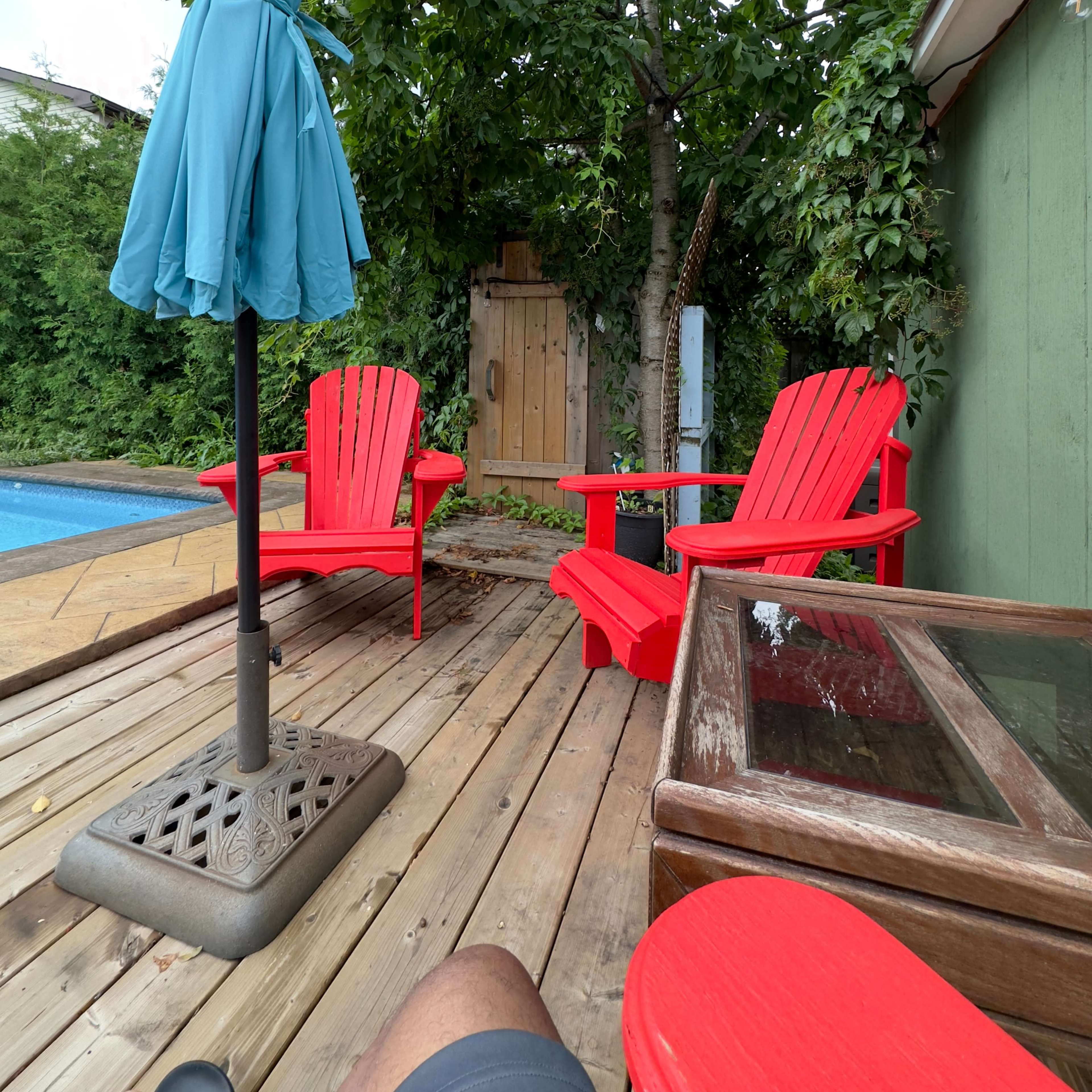 A wooden deck with two red Adirondack chairs, a blue umbrella, and a small shed partially obscured by greenery.