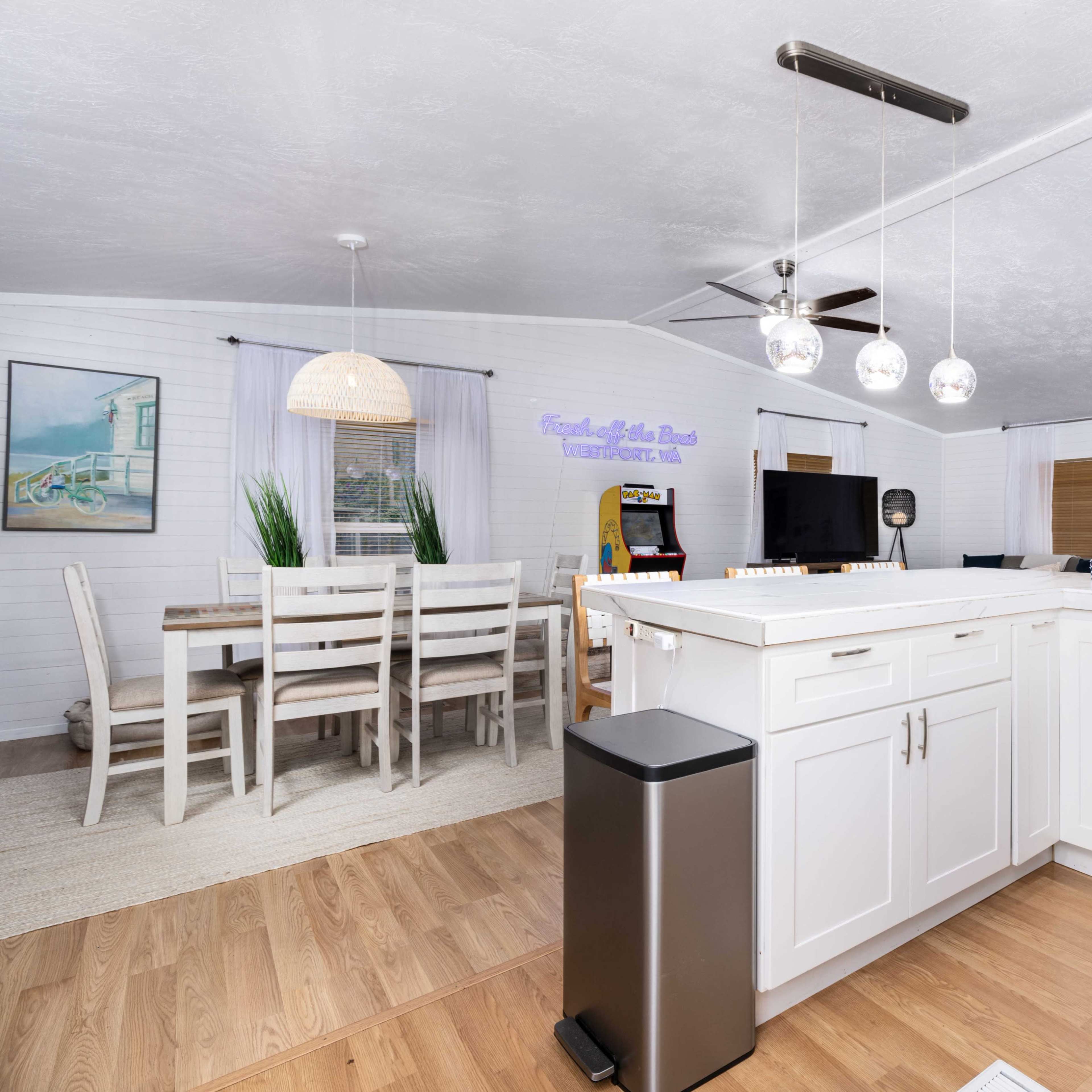 A modern kitchen with white cabinetry and stainless steel appliances, adjacent to a dining area with a wooden table and light fixtures.
