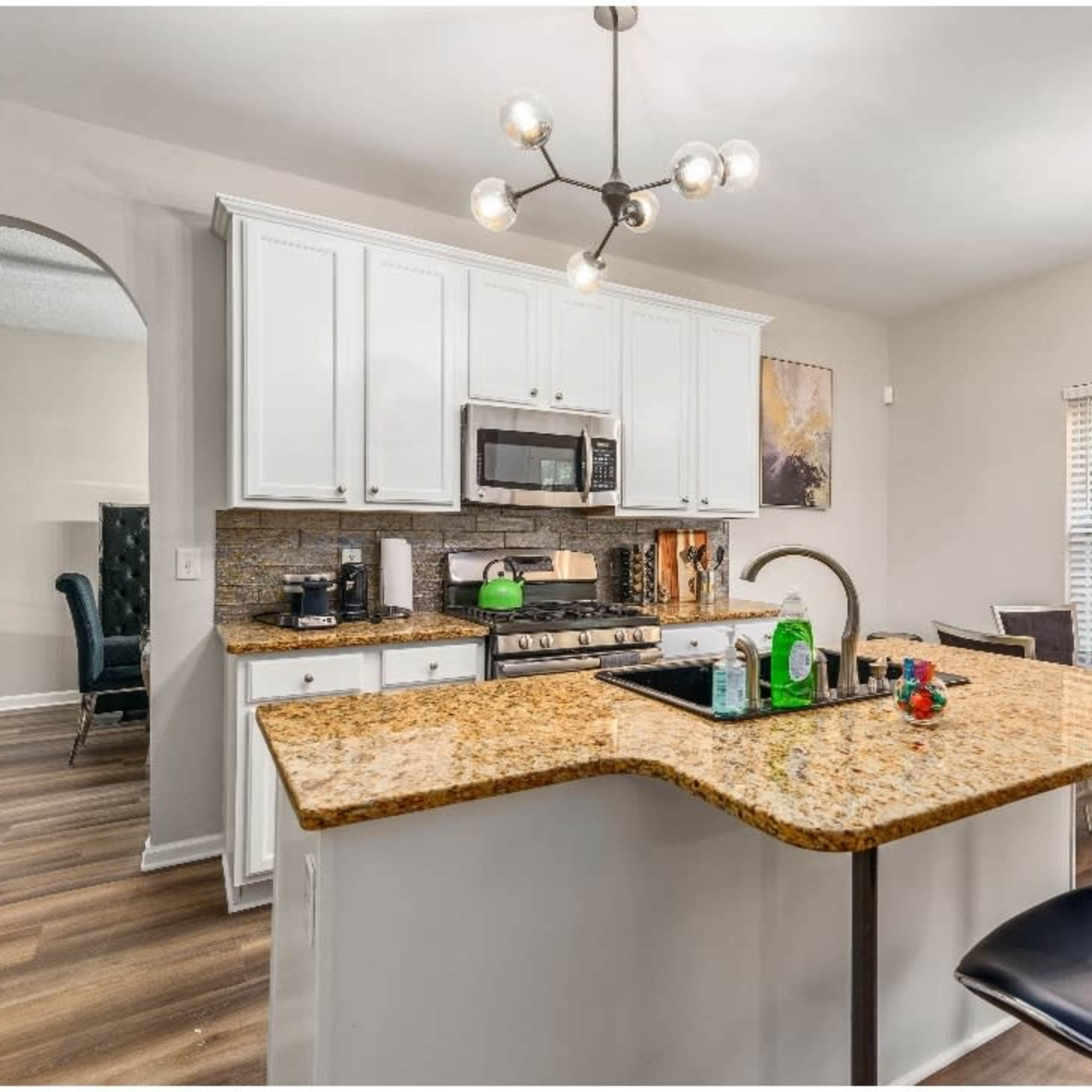 A modern kitchen with white cabinets, a granite countertop, stainless steel appliances, and a dining area featuring a round light fixture.