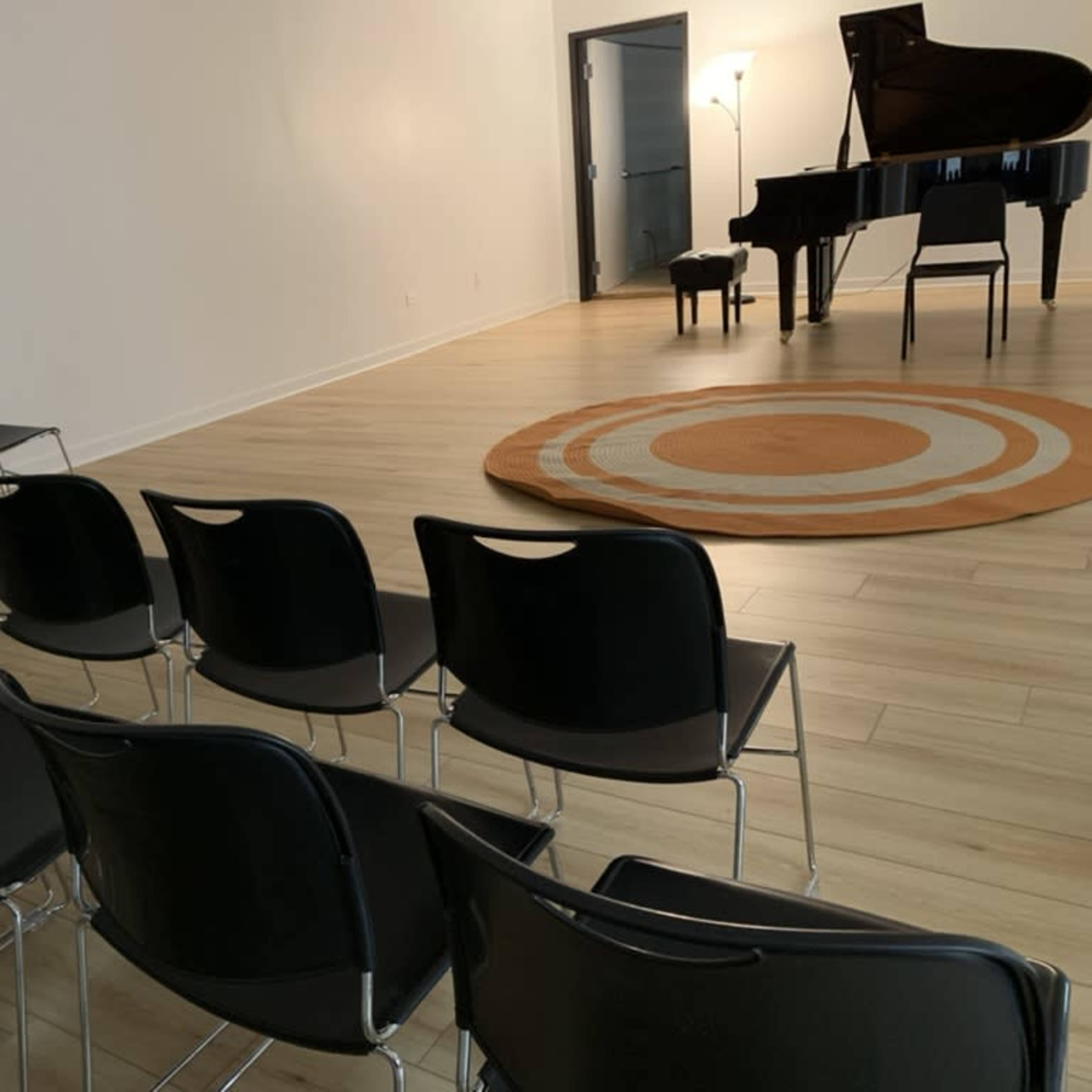 A row of black chairs facing a grand piano on a circular rug in a brightly lit room.