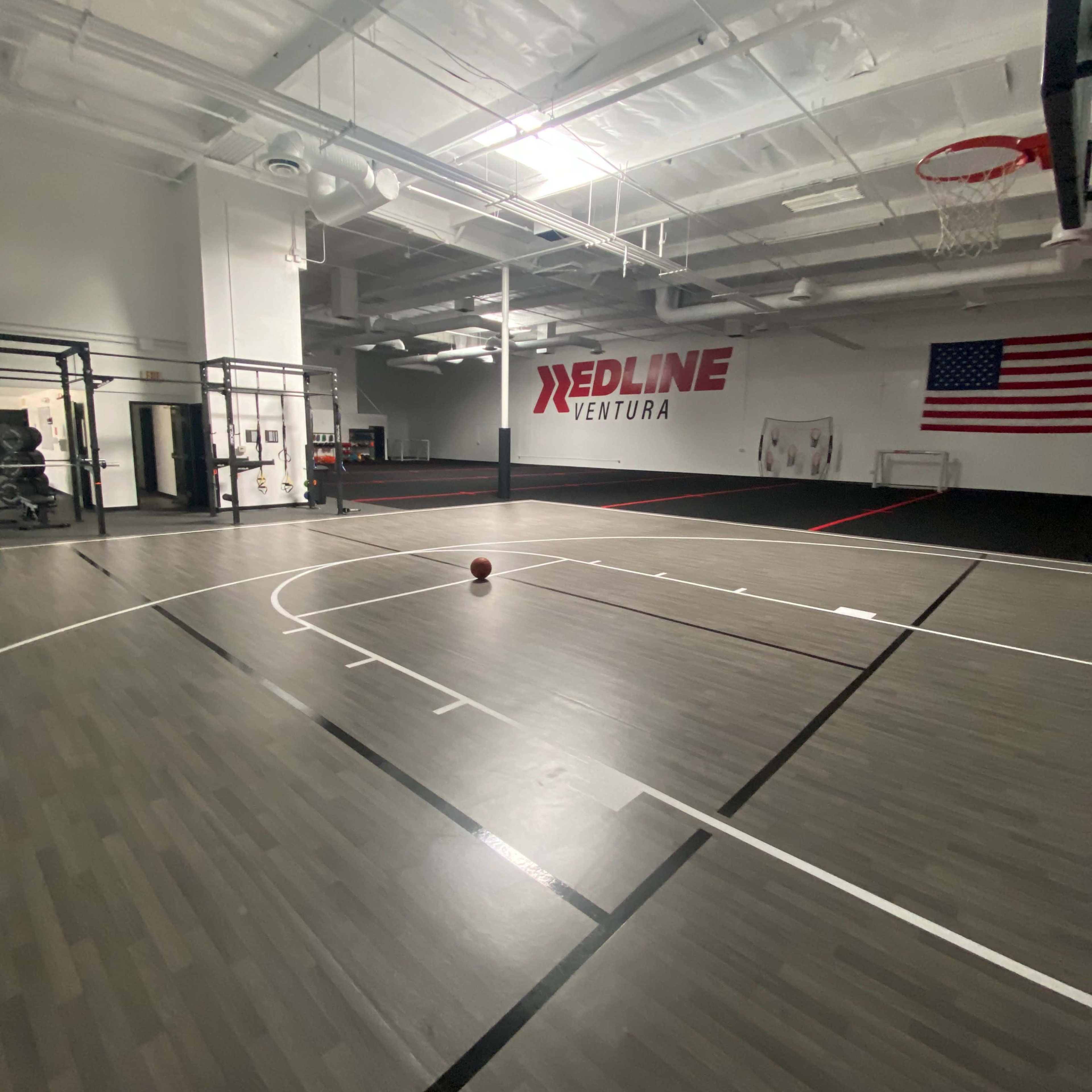 An empty gymnasium with a basketball on the court and a large American flag on the wall.