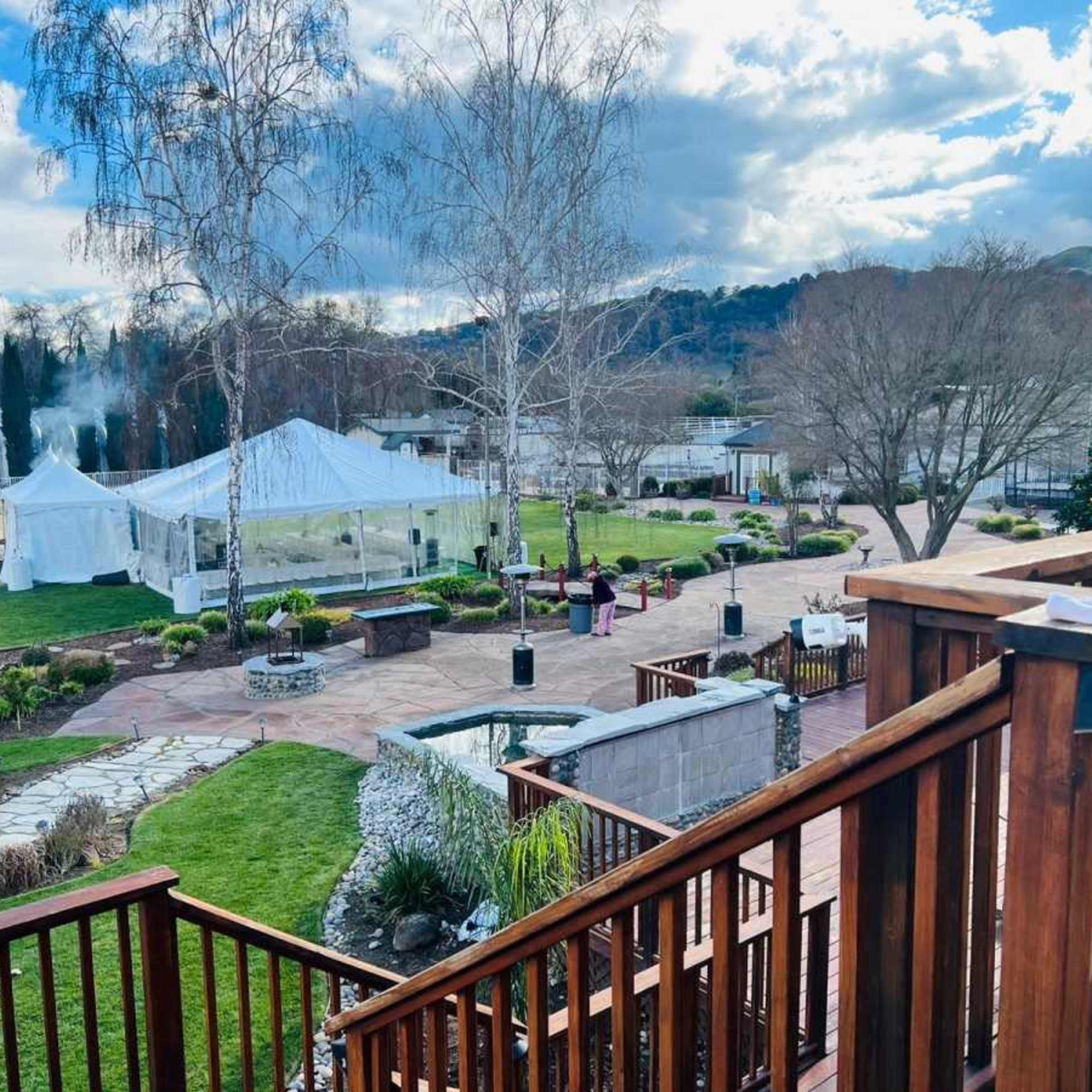 A view from a wooden balcony shows a landscaped outdoor area with a white tent, several seating arrangements, and a stone pathway leading to a garden.