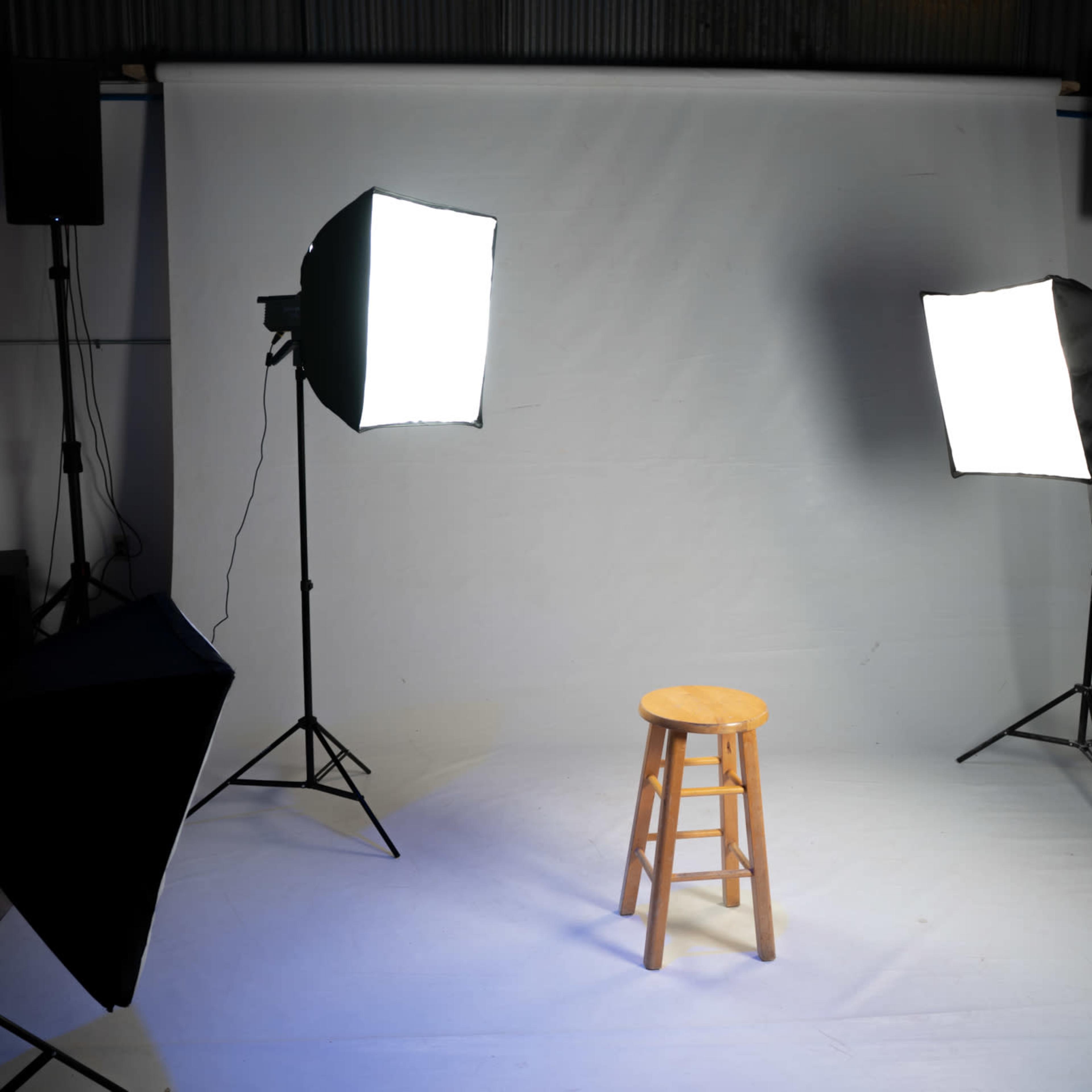 A wooden stool is positioned in front of a plain white backdrop, illuminated by two large softbox lights and flanked by speakers.