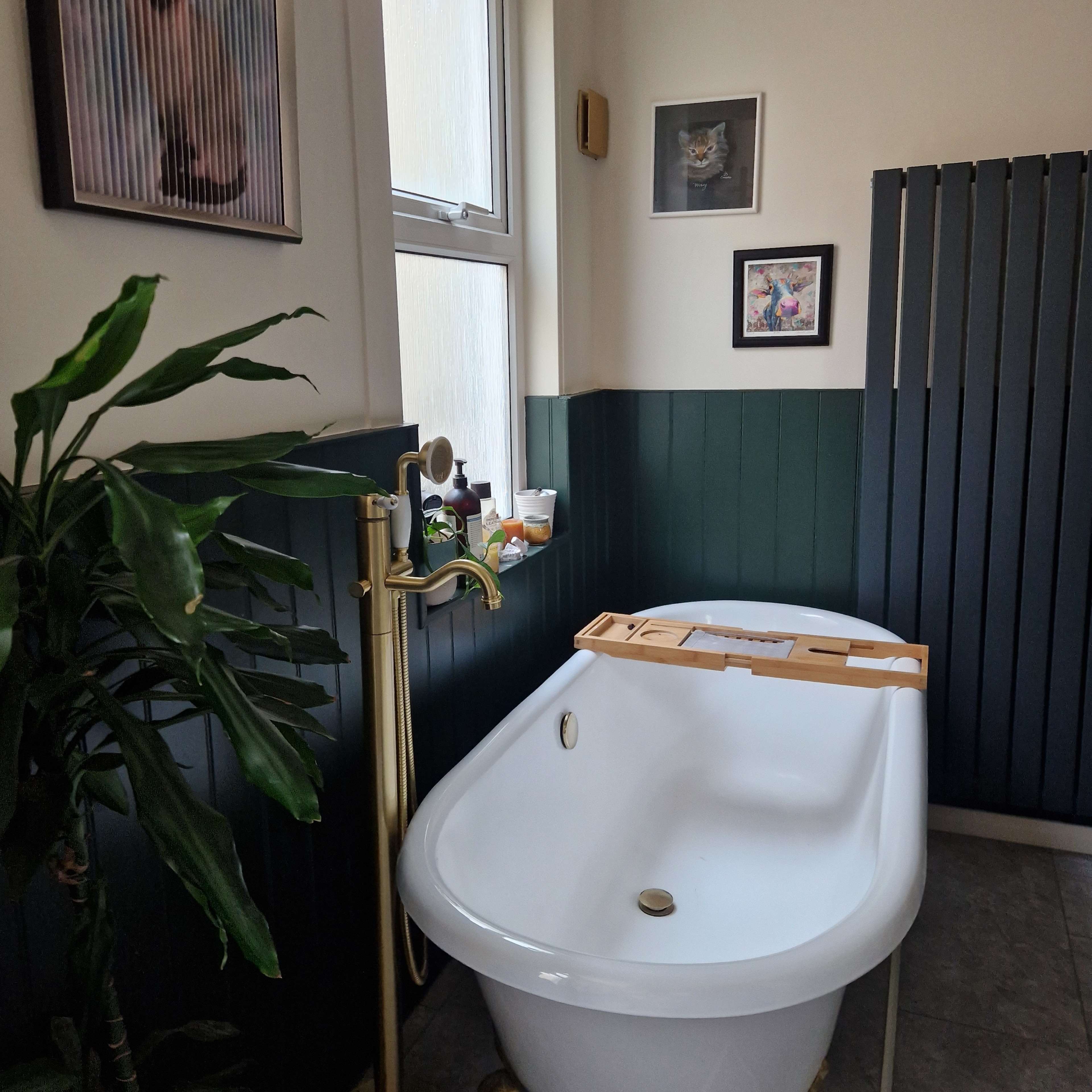 A white freestanding bathtub in a bathroom with green-paneled walls and framed artwork on the walls.