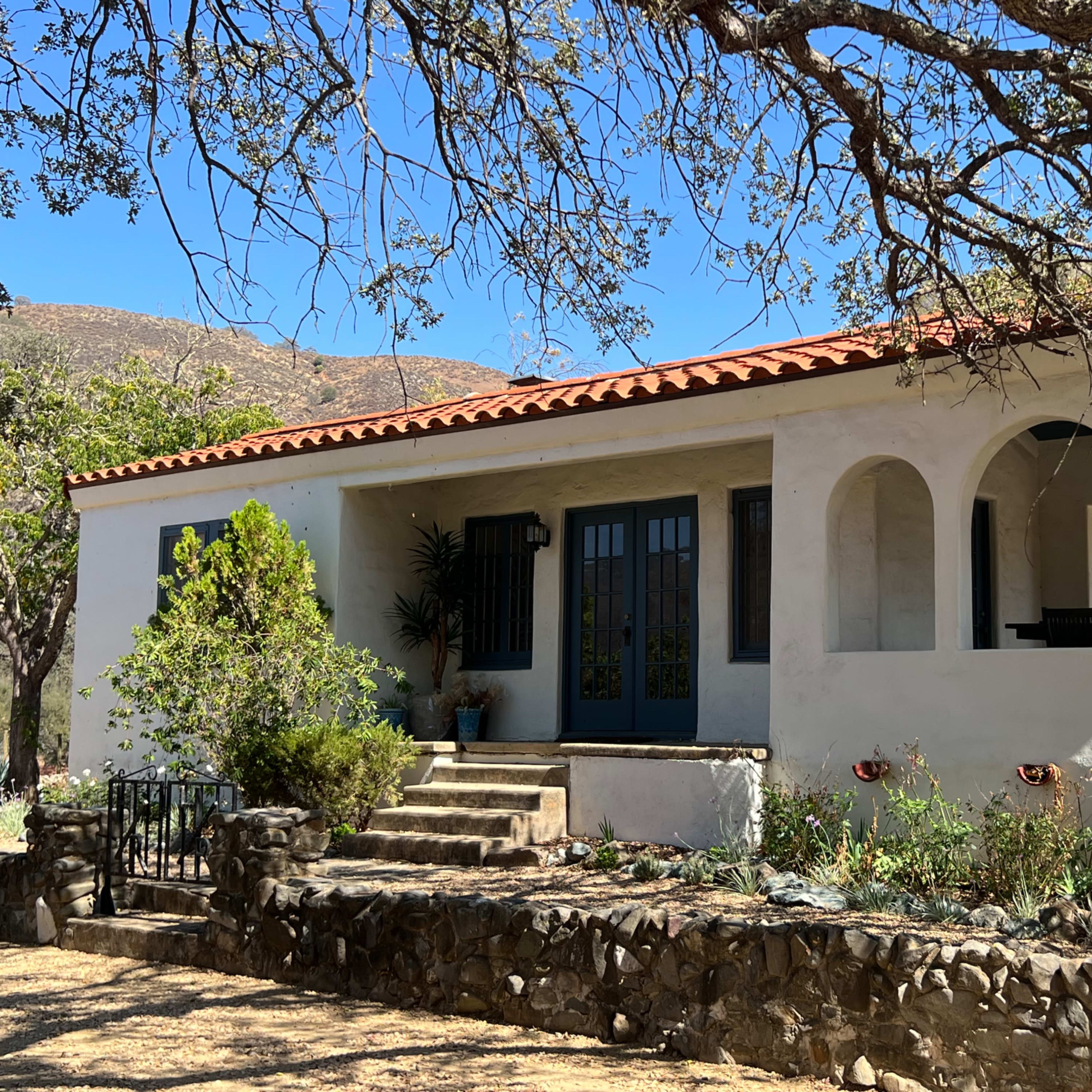 A white, single-story house with a red-tiled roof features arched doorways and a stone walkway surrounded by greenery.