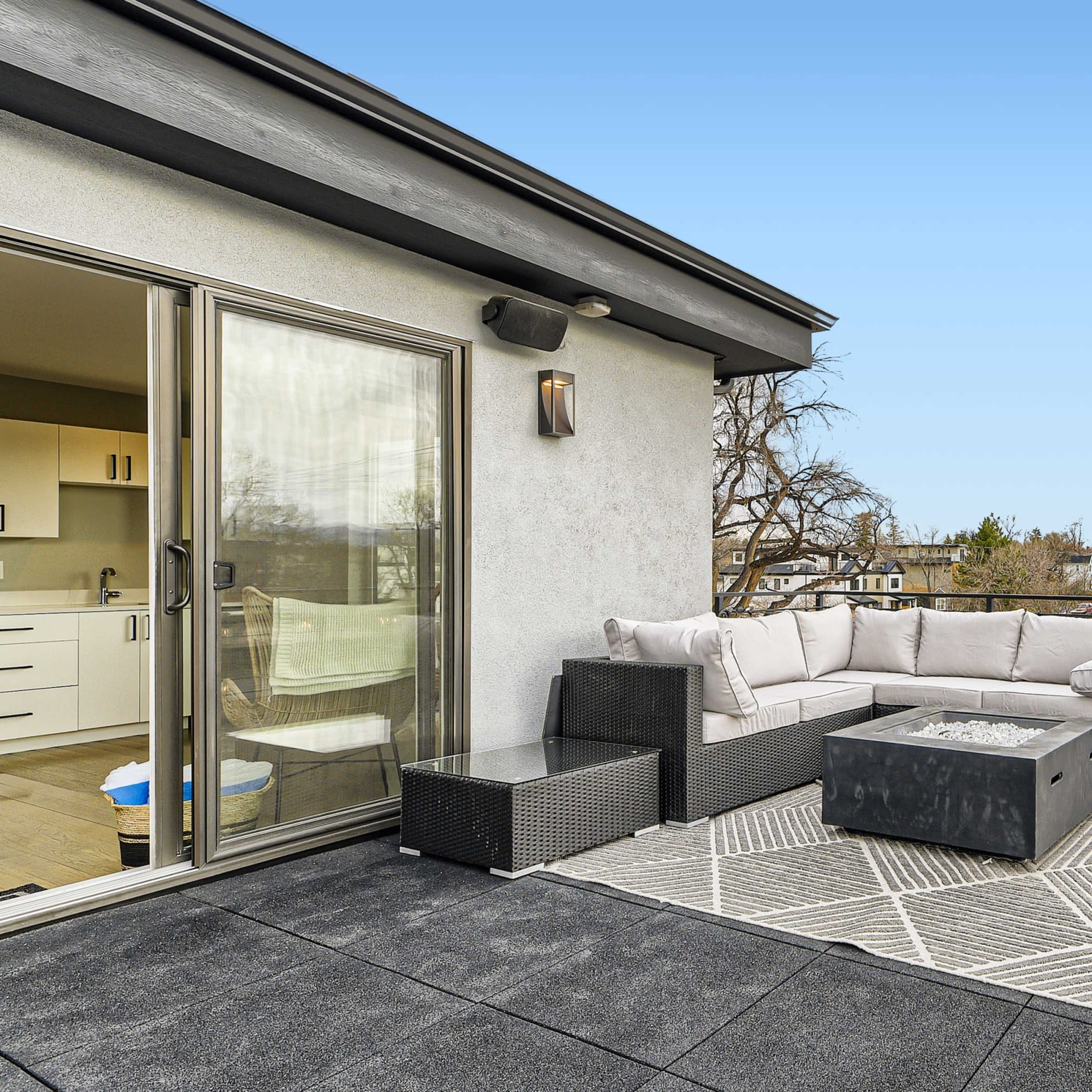 A modern rooftop terrace features a sectional sofa, a fire pit, and a small side table overlooking a neighborhood.
