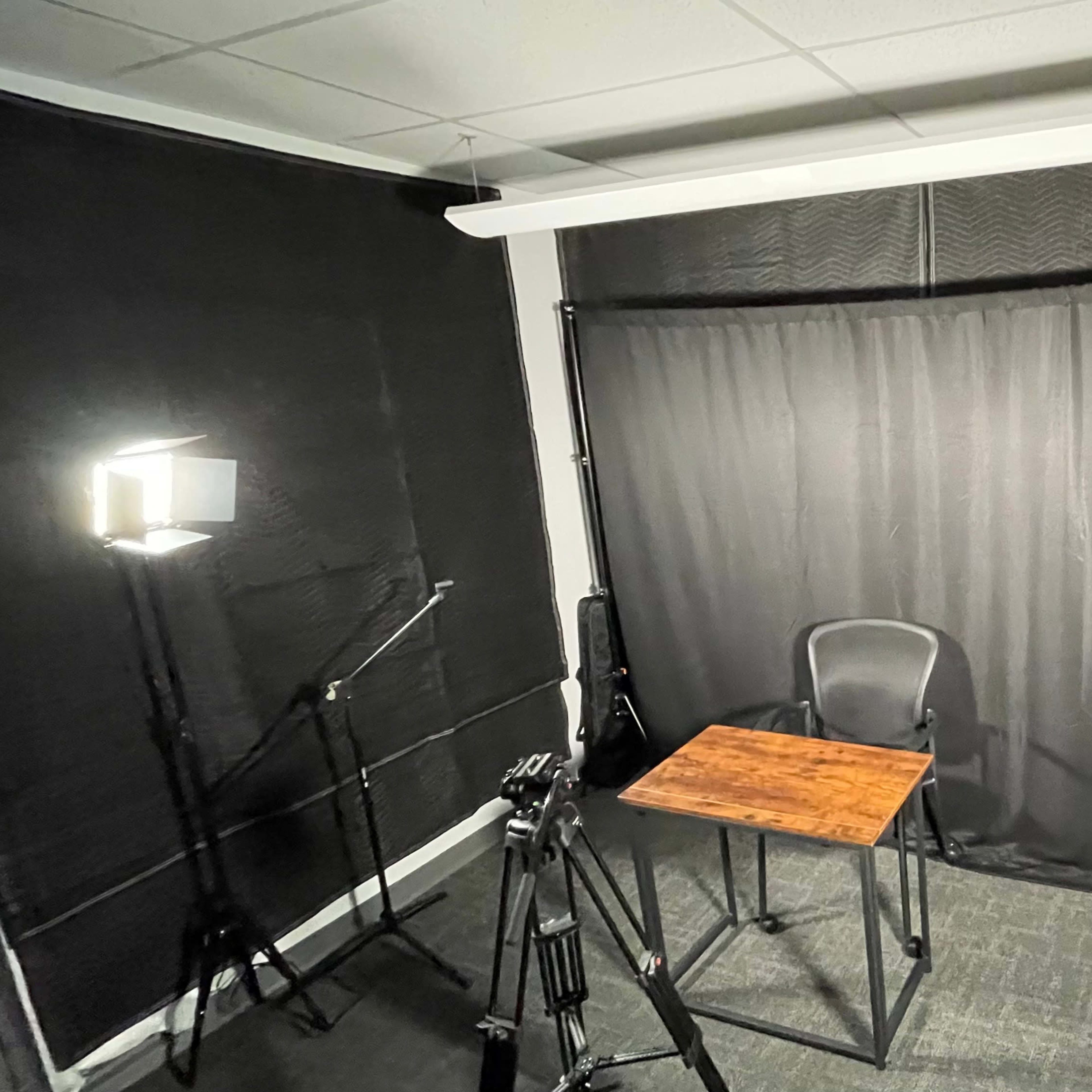 A small recording studio featuring a wooden table and a black backdrop, surrounded by various lighting equipment and tripods.