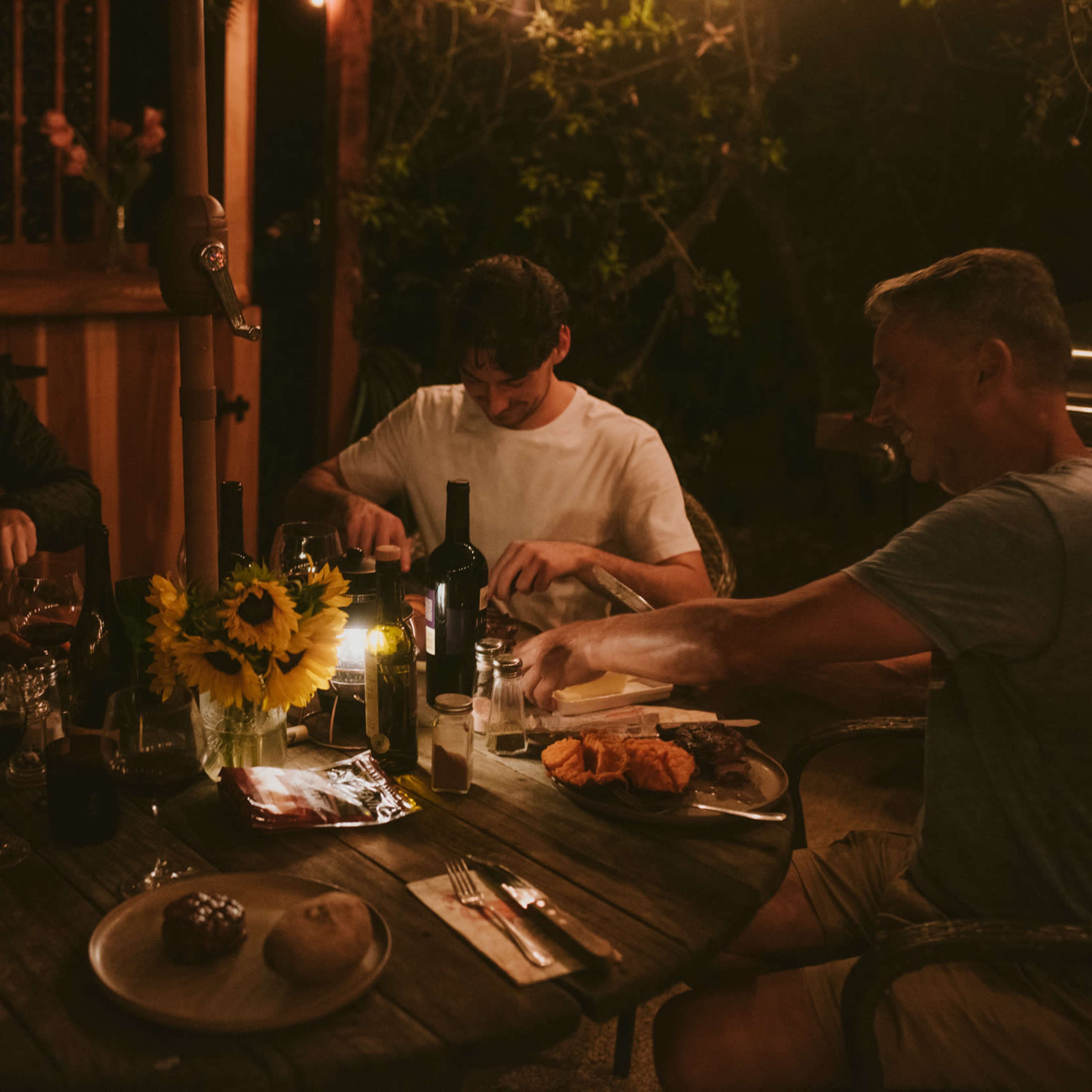 A group of four people are seated around a wooden table, enjoying a dinner with various dishes and drinks under warm lighting.