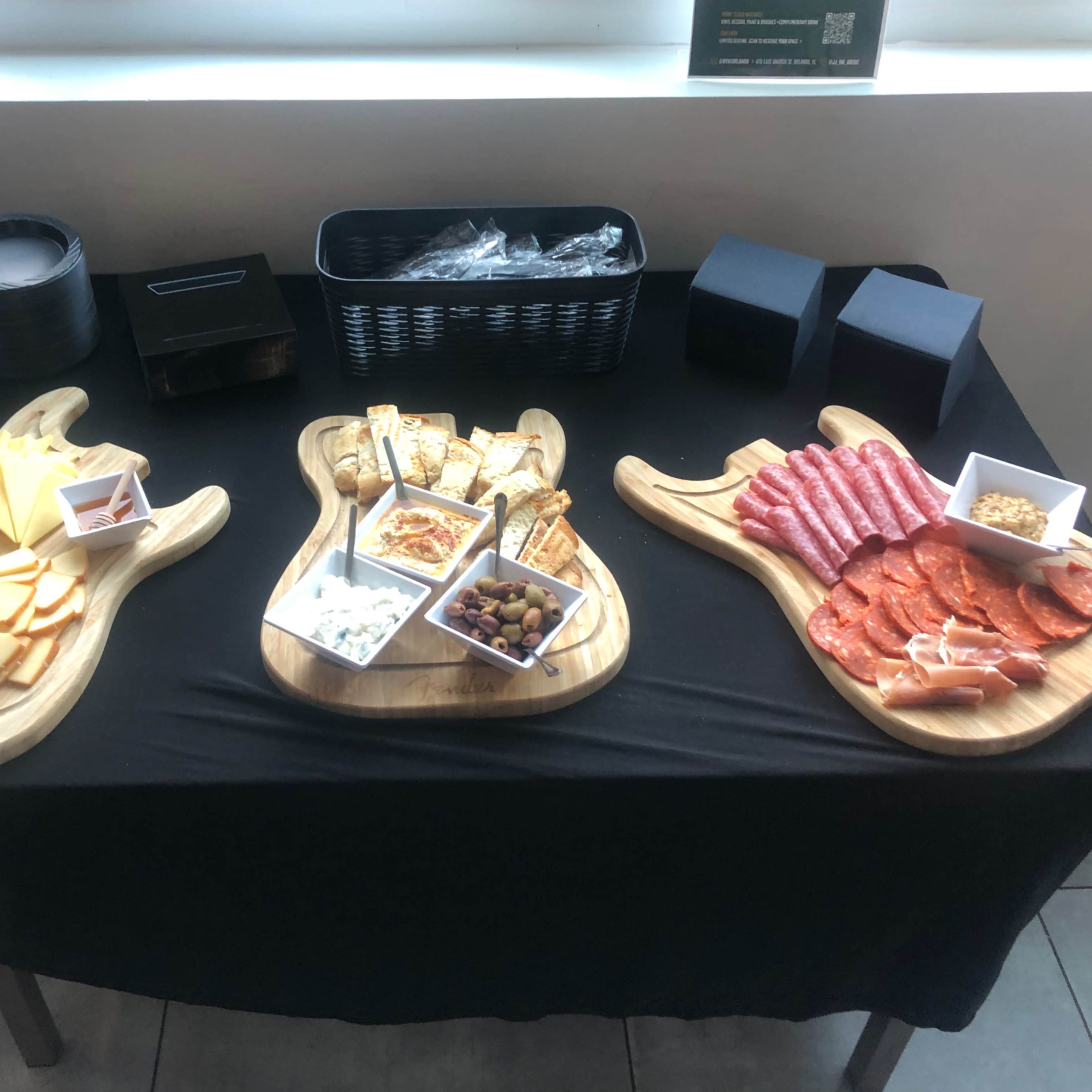A table covered with a black cloth, featuring three wooden boards shaped like guitars, each displaying an assortment of cheeses, meats, fruits, and snacks in small bowls.
