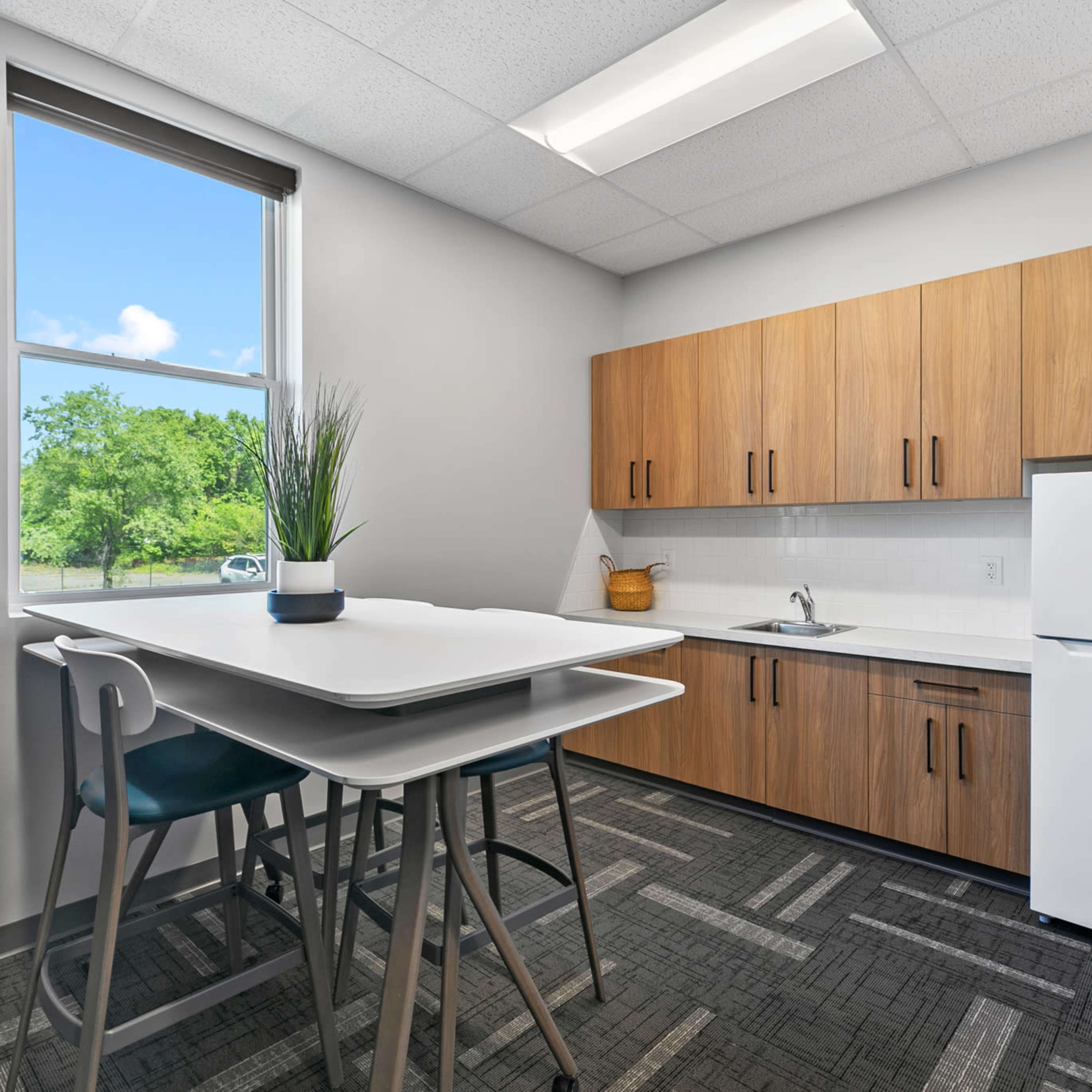 A modern kitchen features wooden cabinets, a white refrigerator, and a tall table with chairs beside a window.