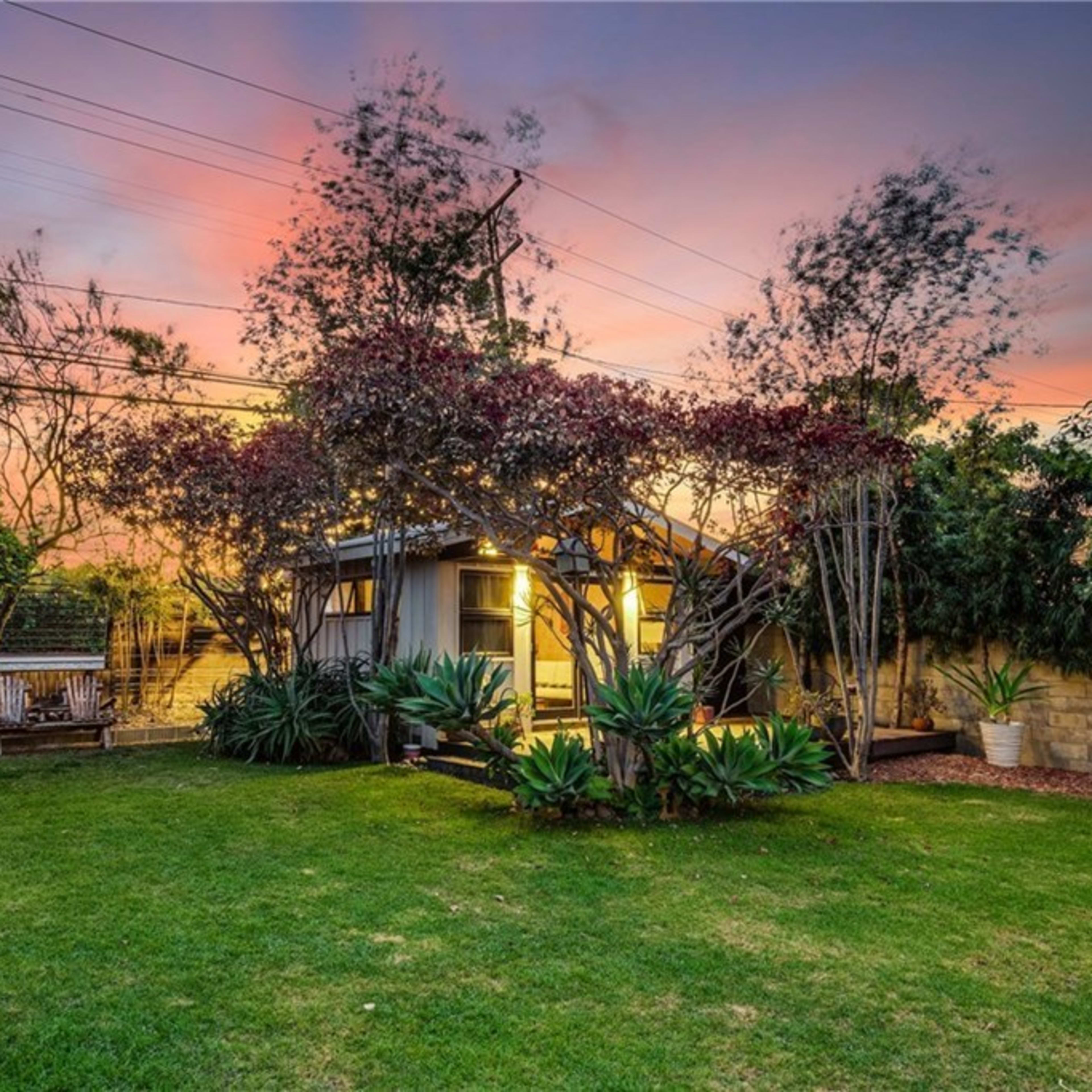 A small house is set in a lush backyard surrounded by trees and plants, with the sky displaying hues of pink and orange at sunset.