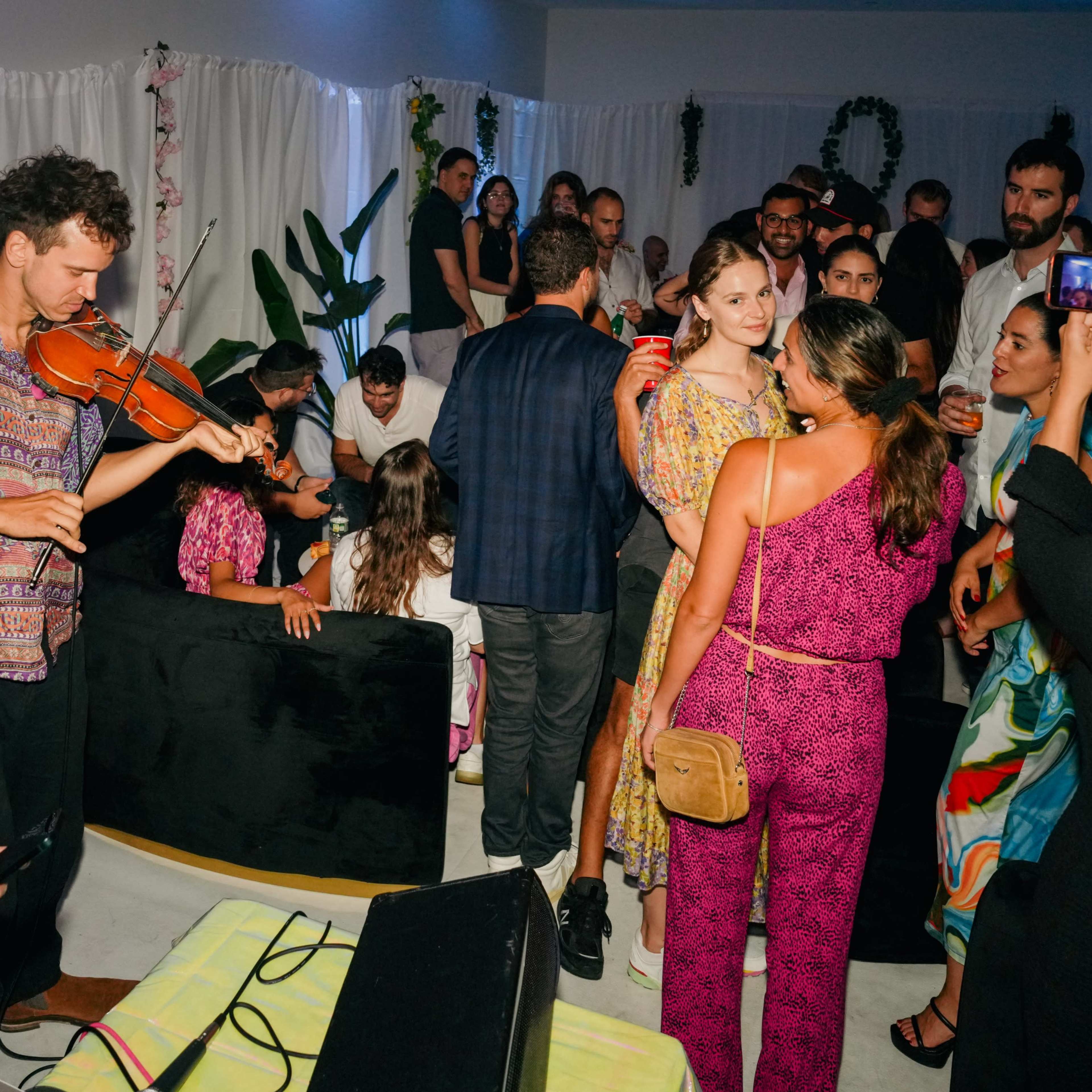 A group of people socialize at a vibrant indoor gathering, with one person playing the violin while others chat and take photos.
