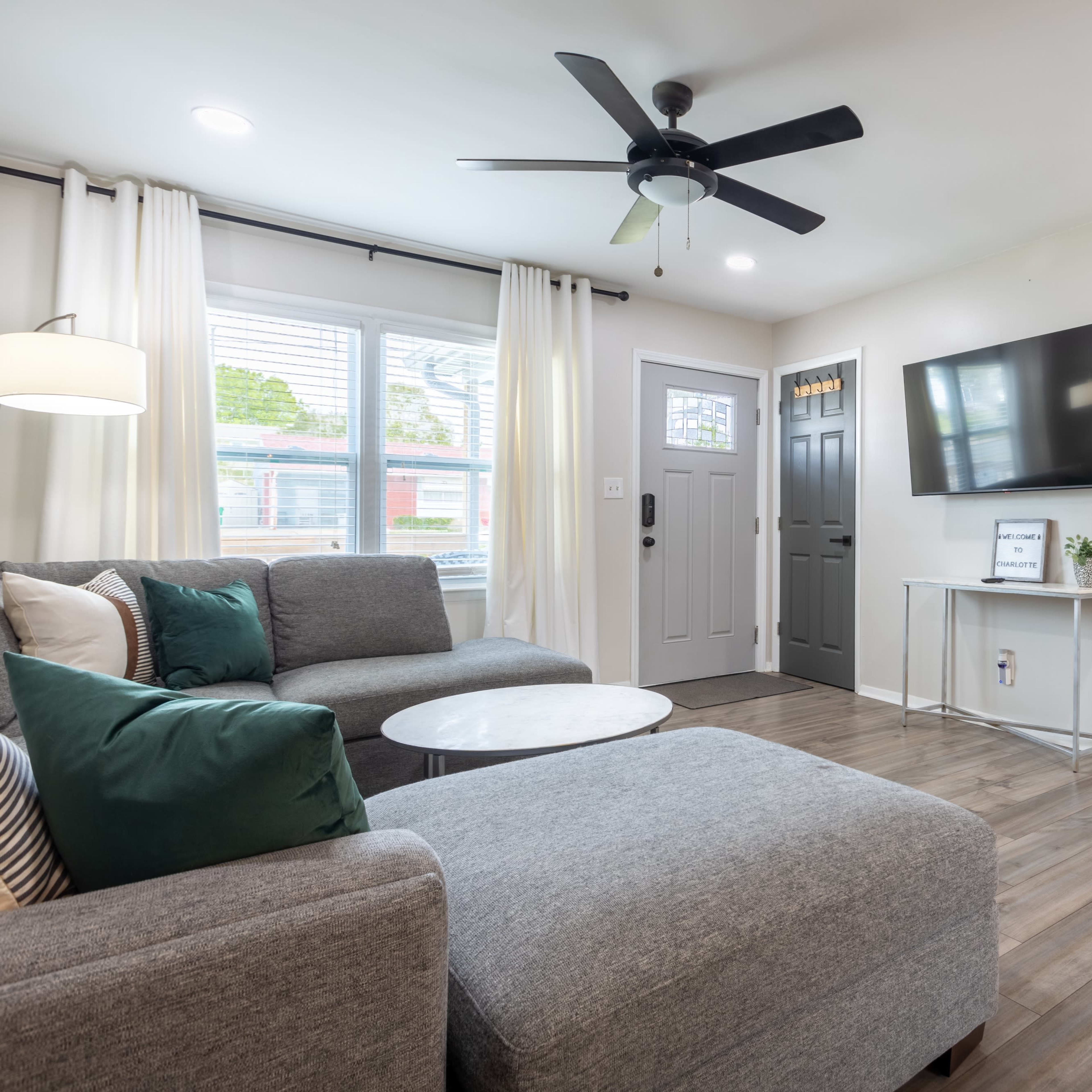 A modern living room featuring a gray sectional sofa, a circular coffee table, a ceiling fan, and a mounted television on a light-colored wall.